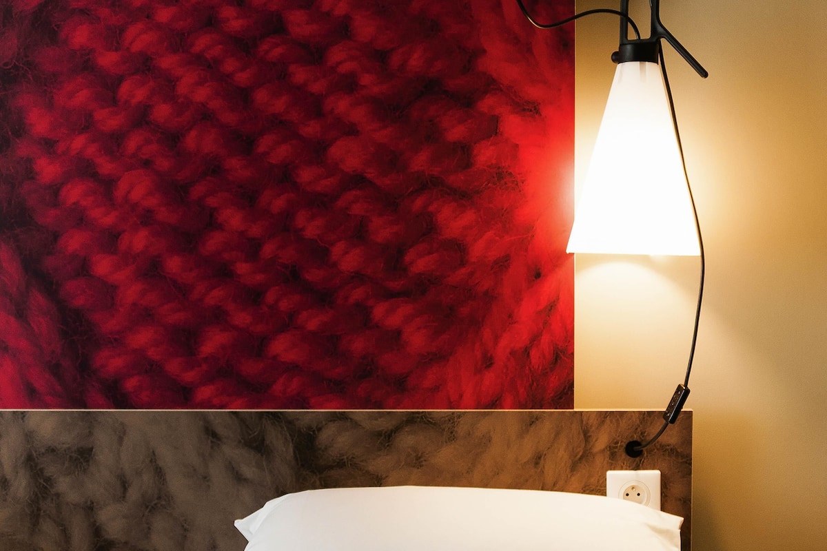 A hotel room with a red textured wall, a white bed, and a bedside table with a lamp.