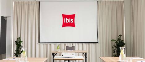 A conference room with a projector screen displaying the 'ibis' logo, a table with a laptop, and two potted plants.