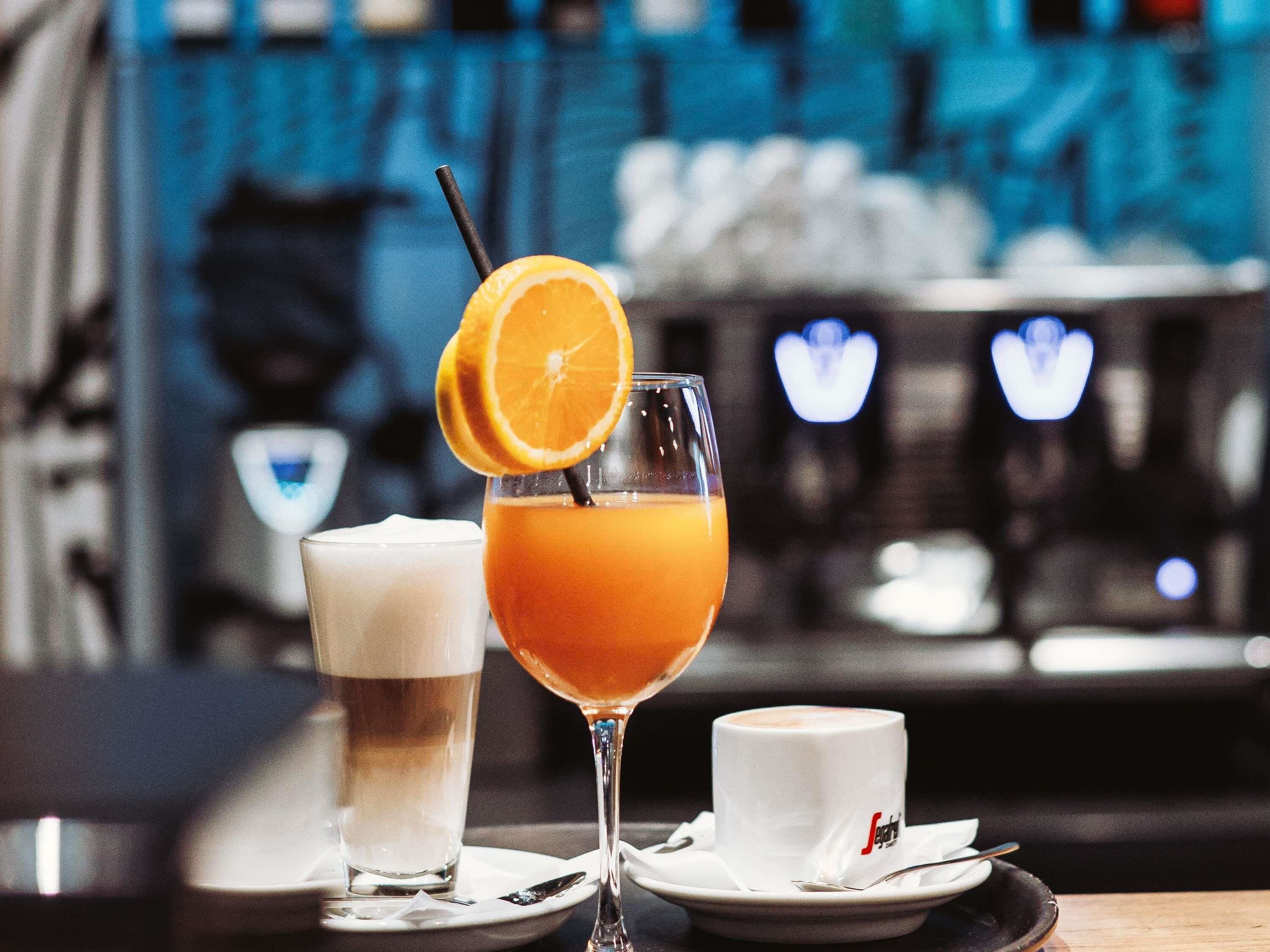 Eine Bar mit einem Glas Orangensaft, einem hohen Glas mit einem hellen Getränk und einer Tasse Kaffee auf einem Tisch.