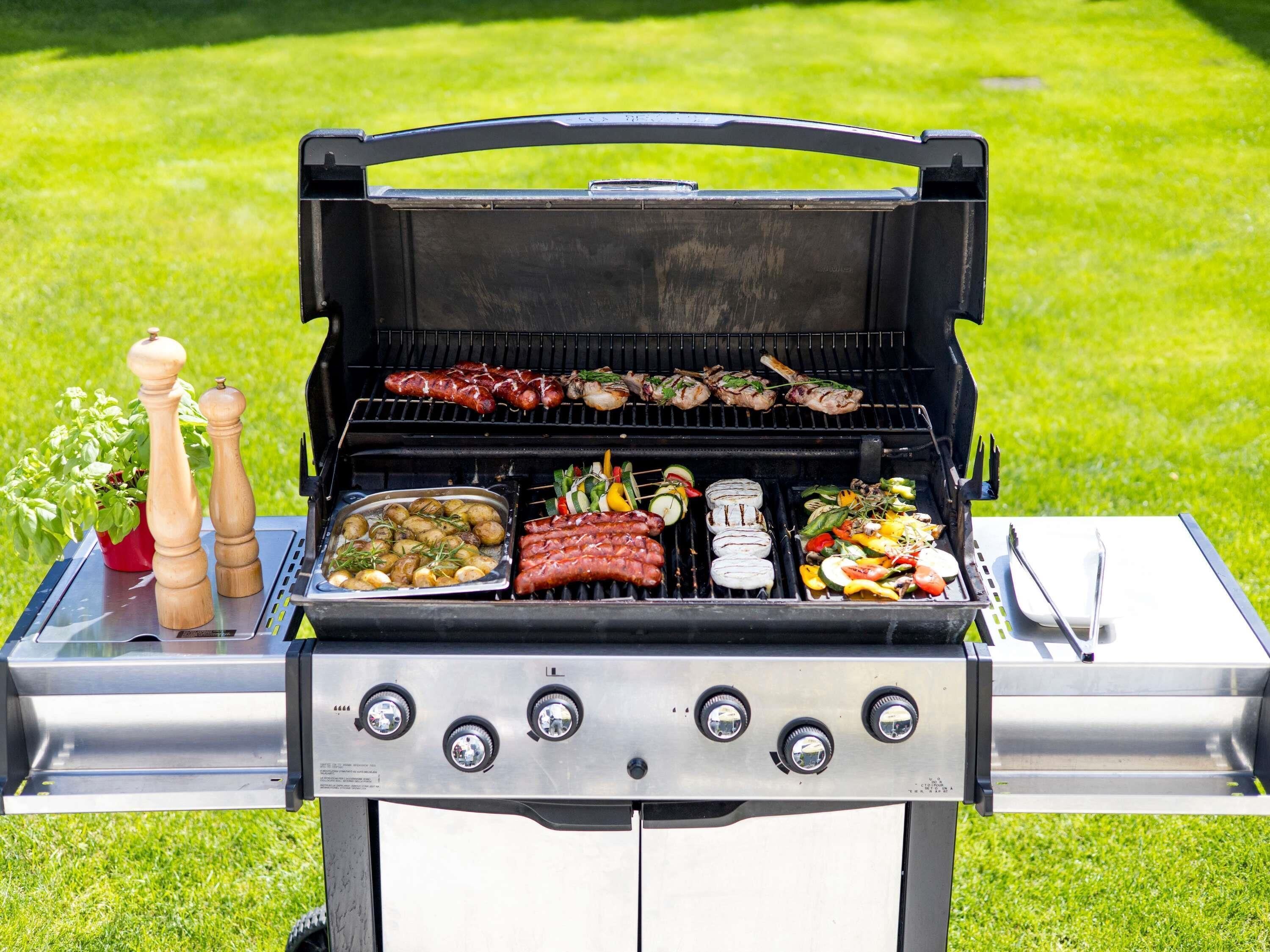 A portable grill with multiple cooking grates, a control panel with knobs, and a stainless steel tray with food.