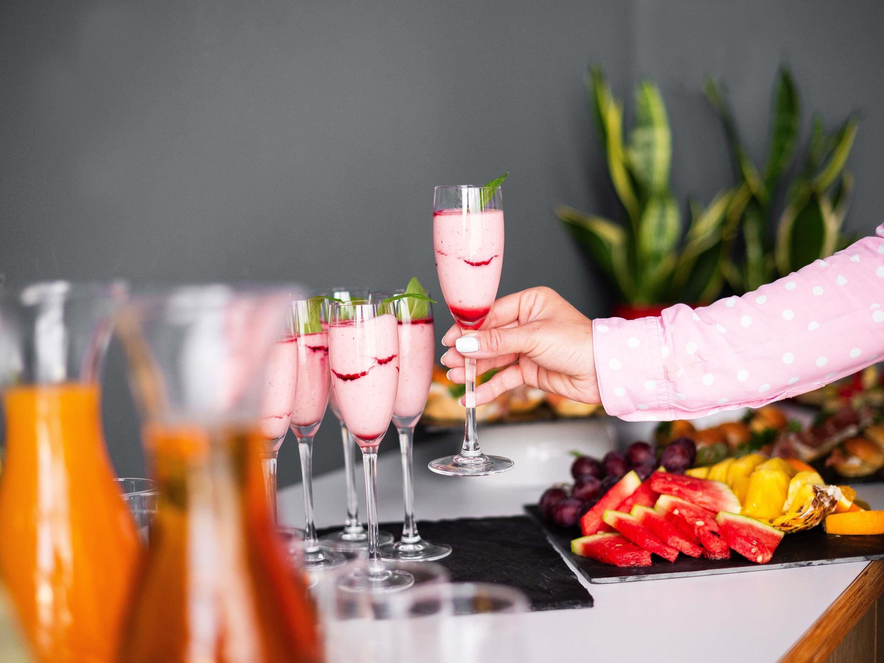 Un bar con varios vasos de una bebida rosada, una mano sosteniendo un vaso y un plato de frutas sobre la barra.