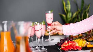 Un bar con varios vasos de una bebida rosada, una mano sosteniendo un vaso y un plato de frutas sobre la barra.