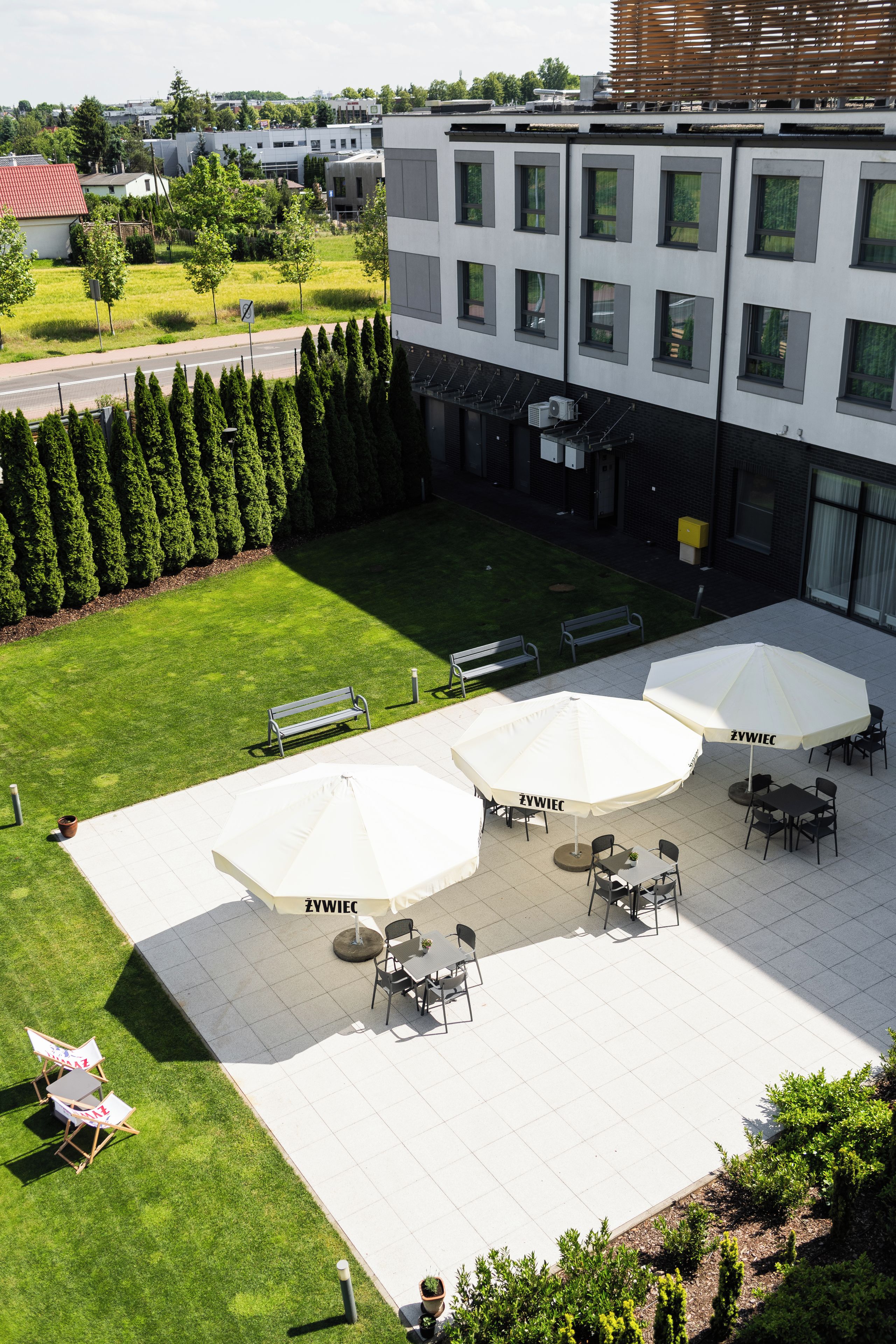 An outdoor seating area with tables and umbrellas, surrounded by a well-maintained lawn and a row of trees.