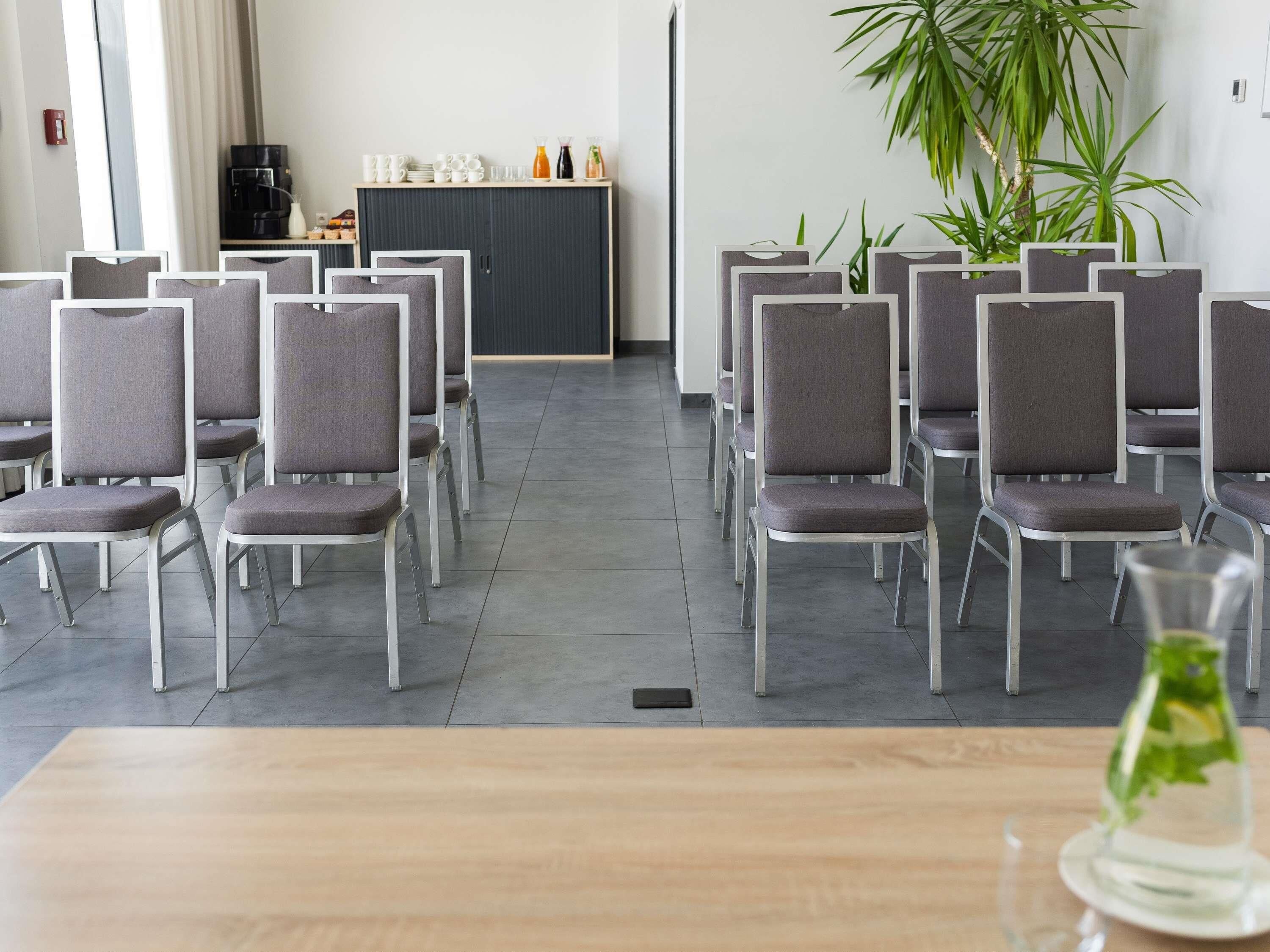 A conference room with rows of grey chairs, a coffee machine, and a plant.