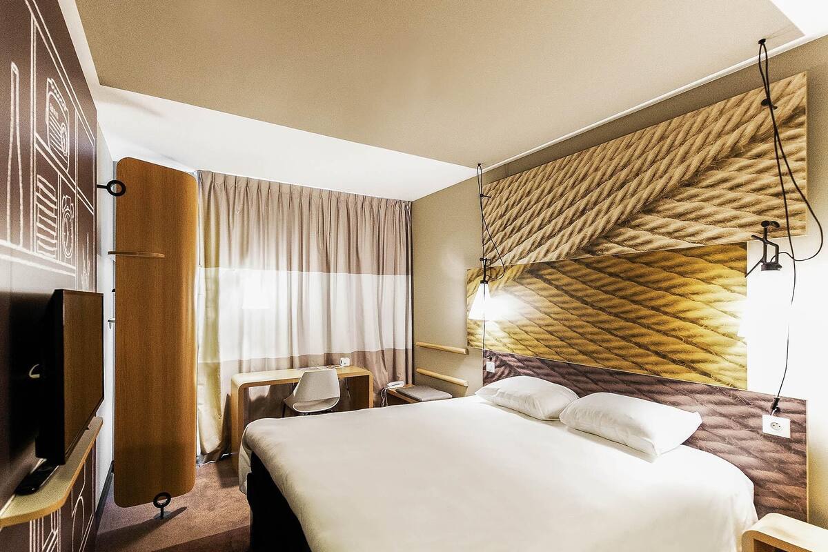 A hotel room with a bed, a desk, a chair, a TV, and a wall with a textured pattern.