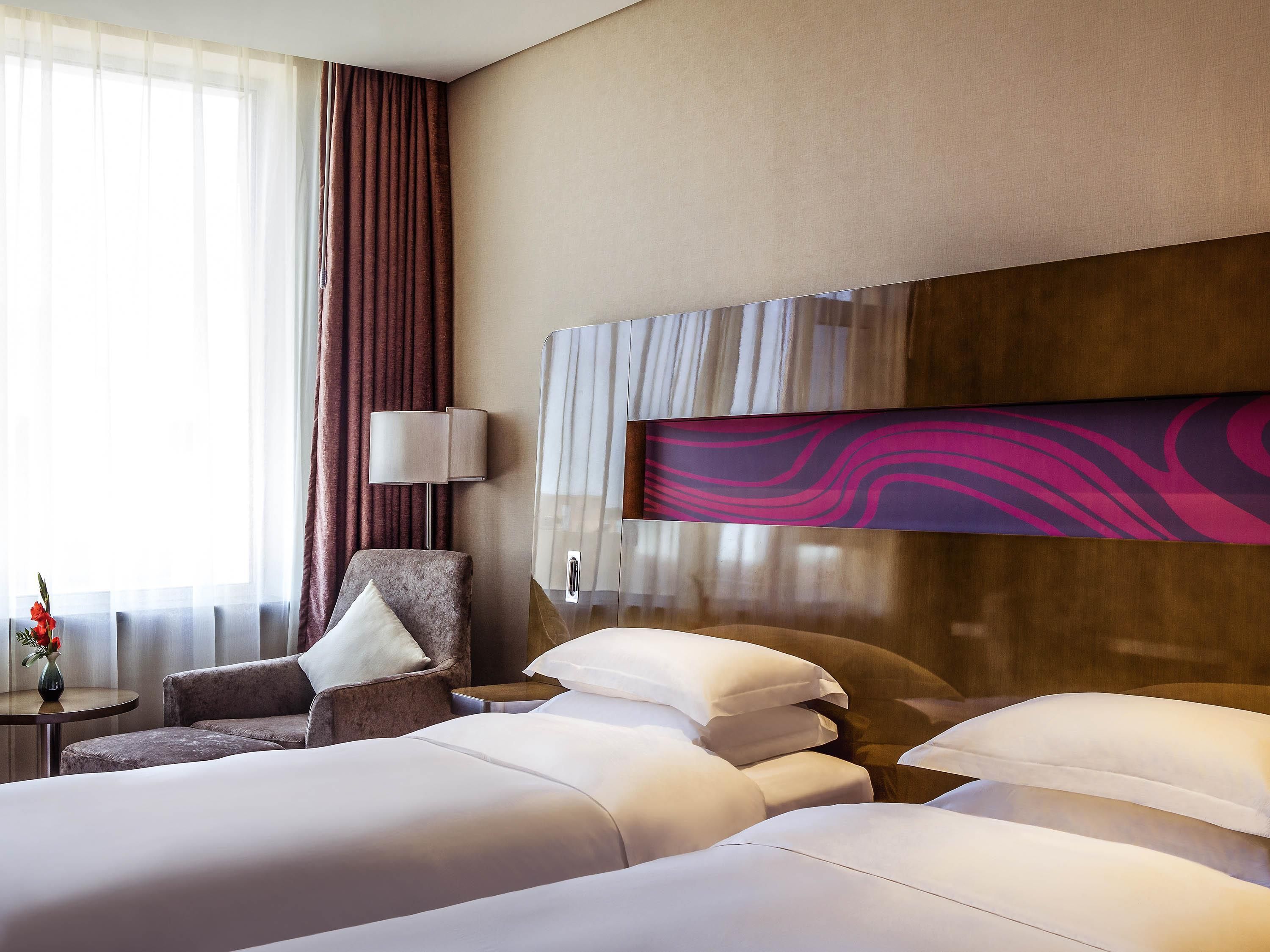 Habitación de hotel con dos camas, una silla, una mesita con un florero y un cuadro abstracto grande en la pared.