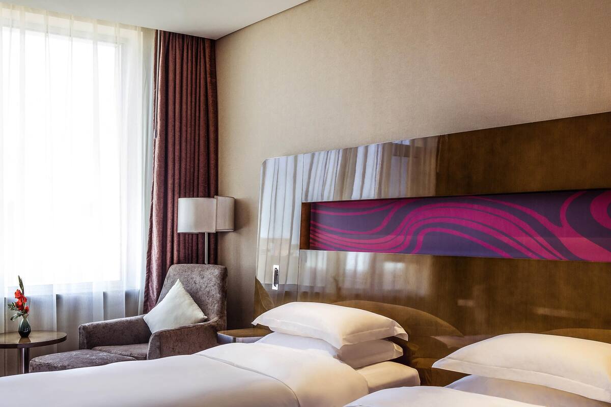 A hotel room with two beds, a chair, a small table with a vase, and a large abstract painting on the wall.
