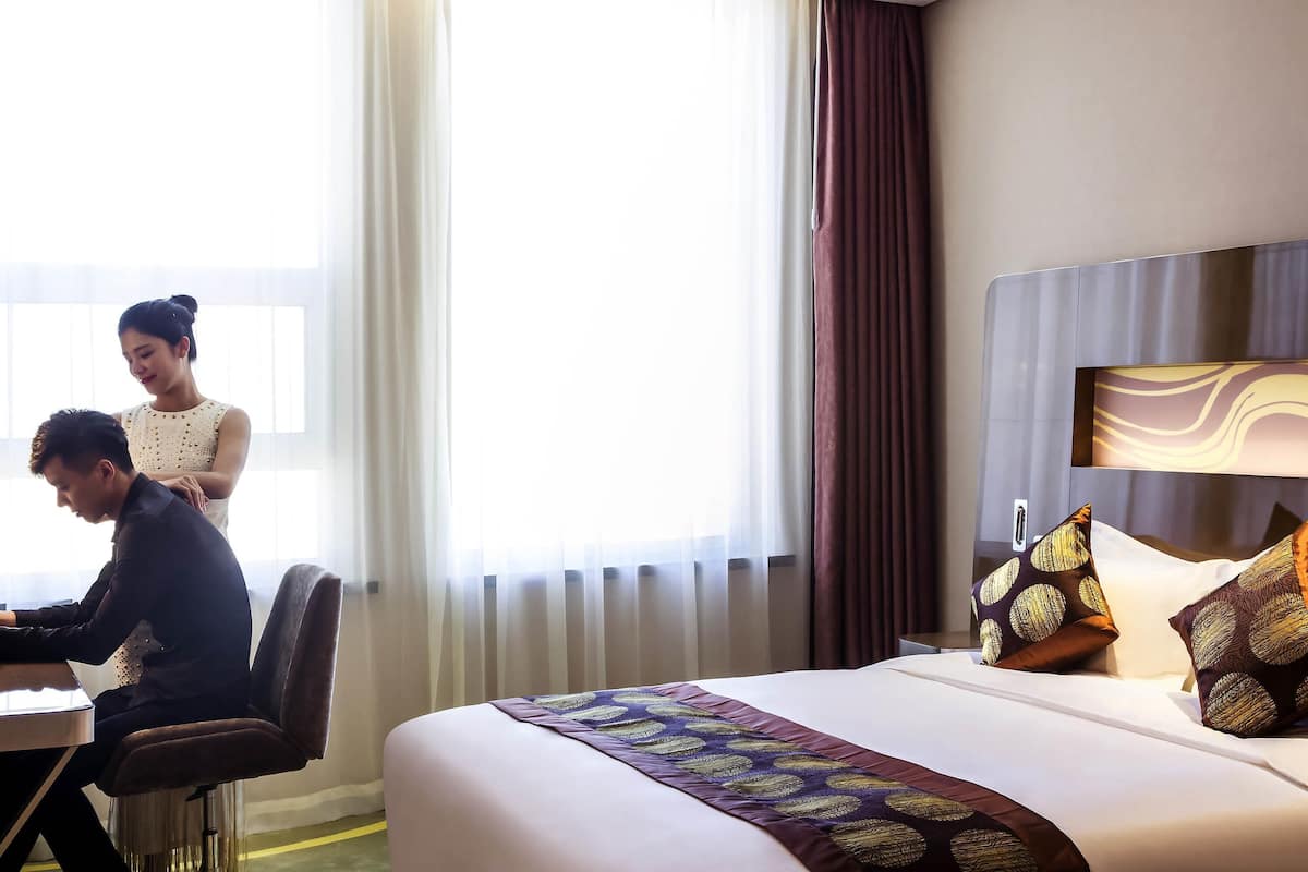 A hotel room with a bed, a desk, and a chair. There is a window with curtains, a wall-mounted lamp, and decorative pillows on the bed.