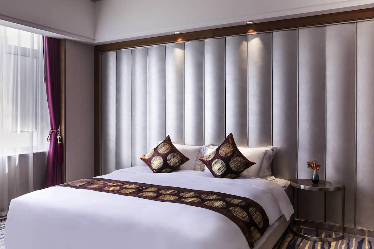 A hotel room with a large bed, a headboard with vertical panels, a bedside table with a vase, and a window with sheer curtains.