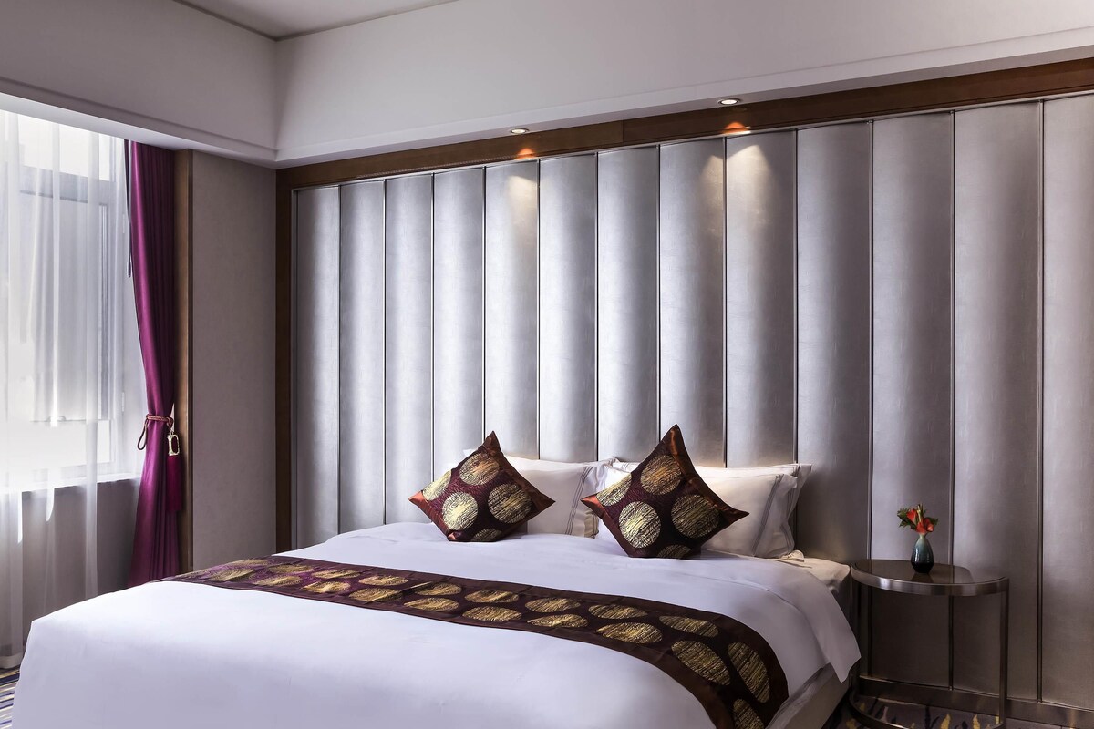 Quarto de hotel com uma cama grande, cabeceira com painéis verticais, mesa de cabeceira com um vaso e janela com cortinas leves.