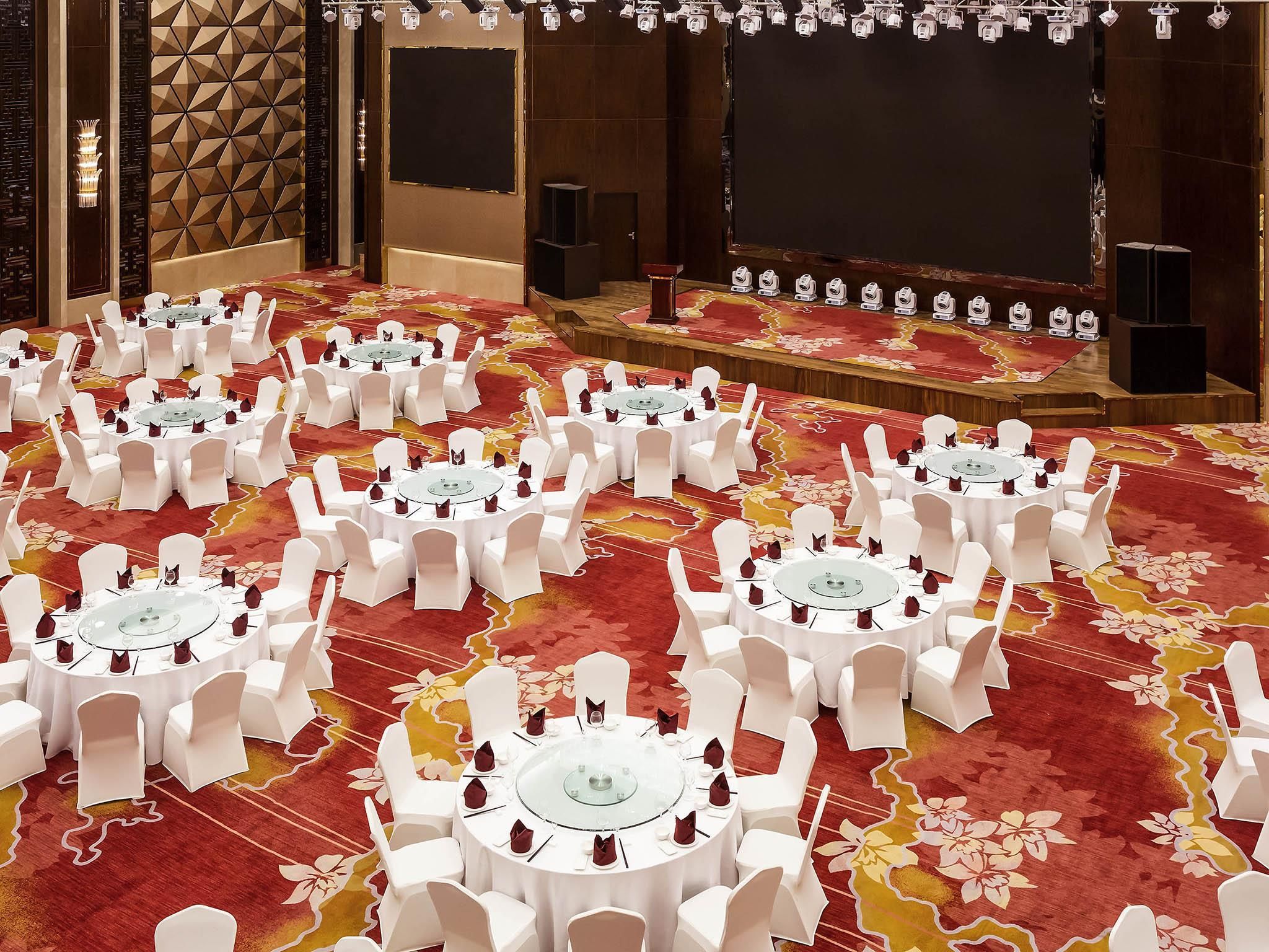 A conference room with multiple round tables, white chairs, and a stage with speakers.