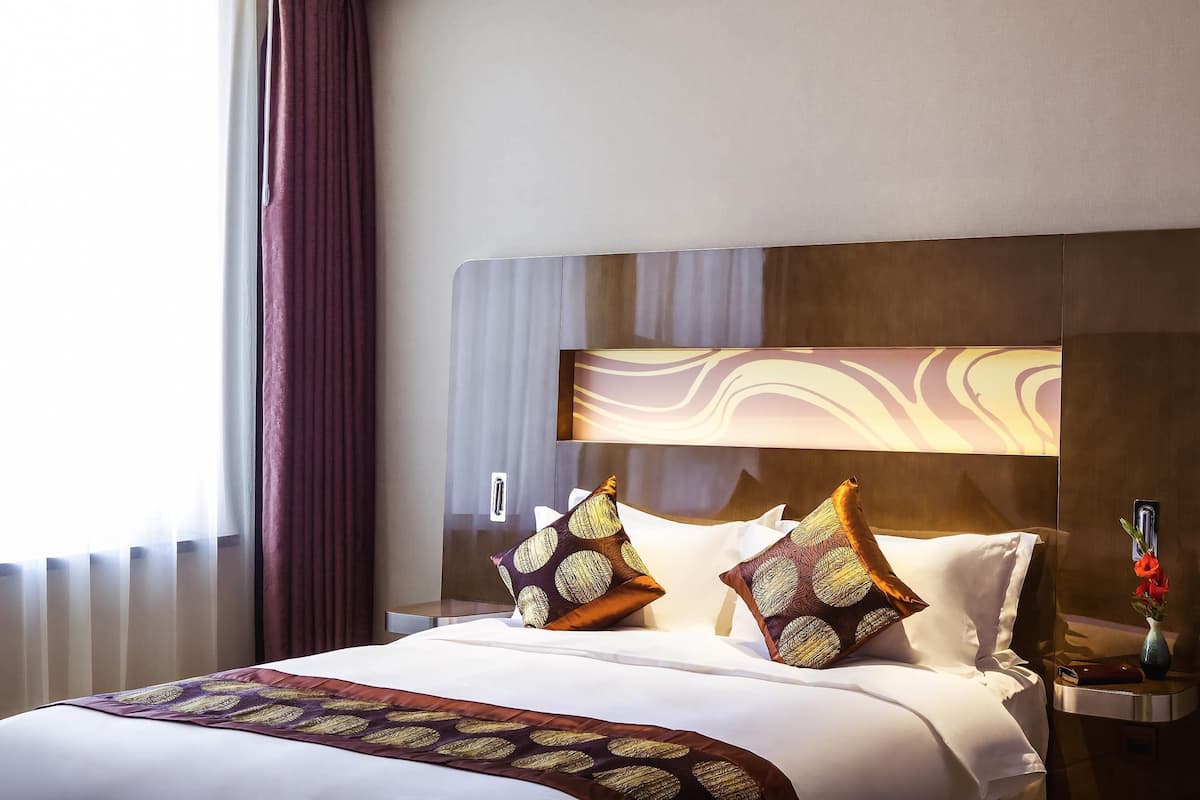 A hotel room with a large bed, a nightstand, a vase with flowers, and a window with curtains.