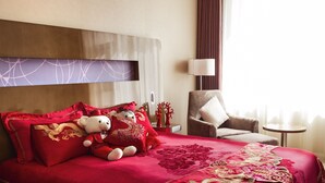 Une chambre d’hôtel avec un grand lit, des oreillers rouges et dorés, un jouet en peluche, une chaise, une petite table et une lampe.