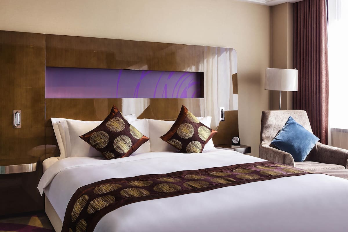 A hotel room with a large bed, a headboard with a built-in TV, a side table, a chair, and a lamp.