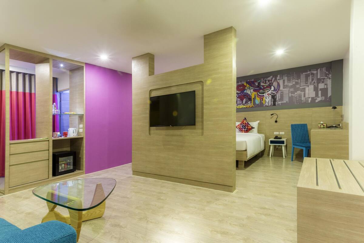 A modern hotel room with a bed, a TV mounted on a wooden wall, a glass coffee table, a blue chair, and a desk with a lamp.