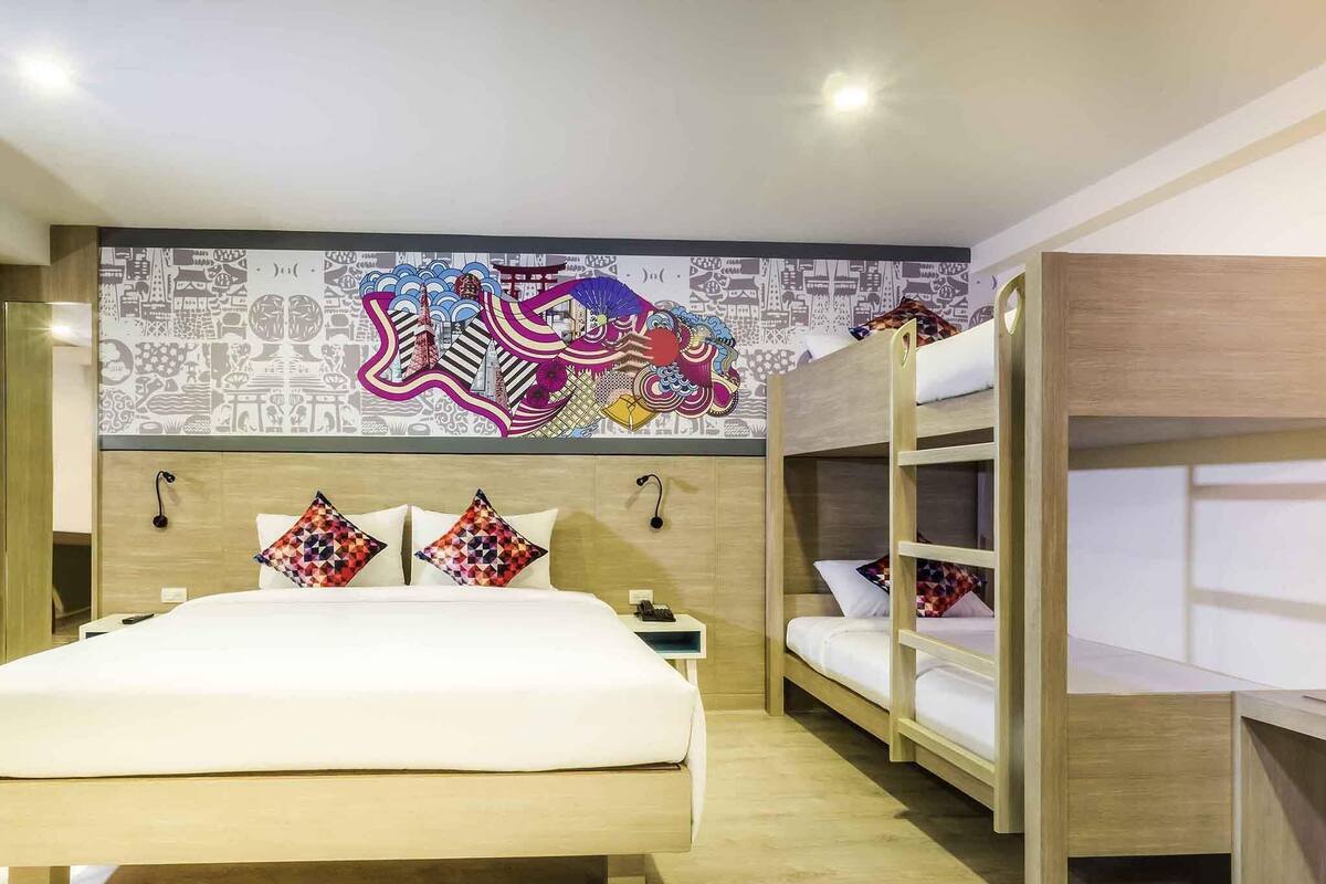 Habitación de hotel con una cama grande, dos camas literas, un mural colorido y un cabecero de madera.