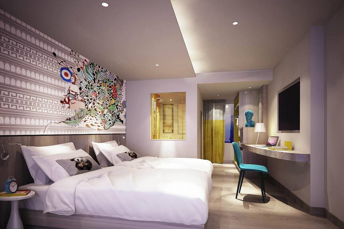 A modern hotel room with a large bed, a desk with a chair, a TV, and a decorative wall.
