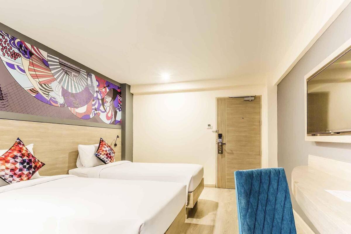 Habitación de hotel con dos camas, un sillón azul, un espejo grande y un cuadro abstracto colorido en la pared.