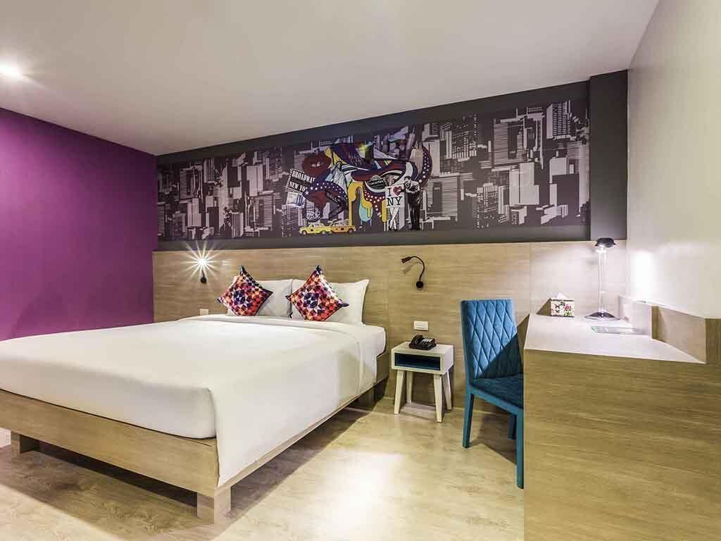 Ein Hotelzimmer mit einem großen Bett, einem kleinen Nachttisch, einem Sessel, einer Wandleuchte und einem Stadtbild-Wandgemälde.