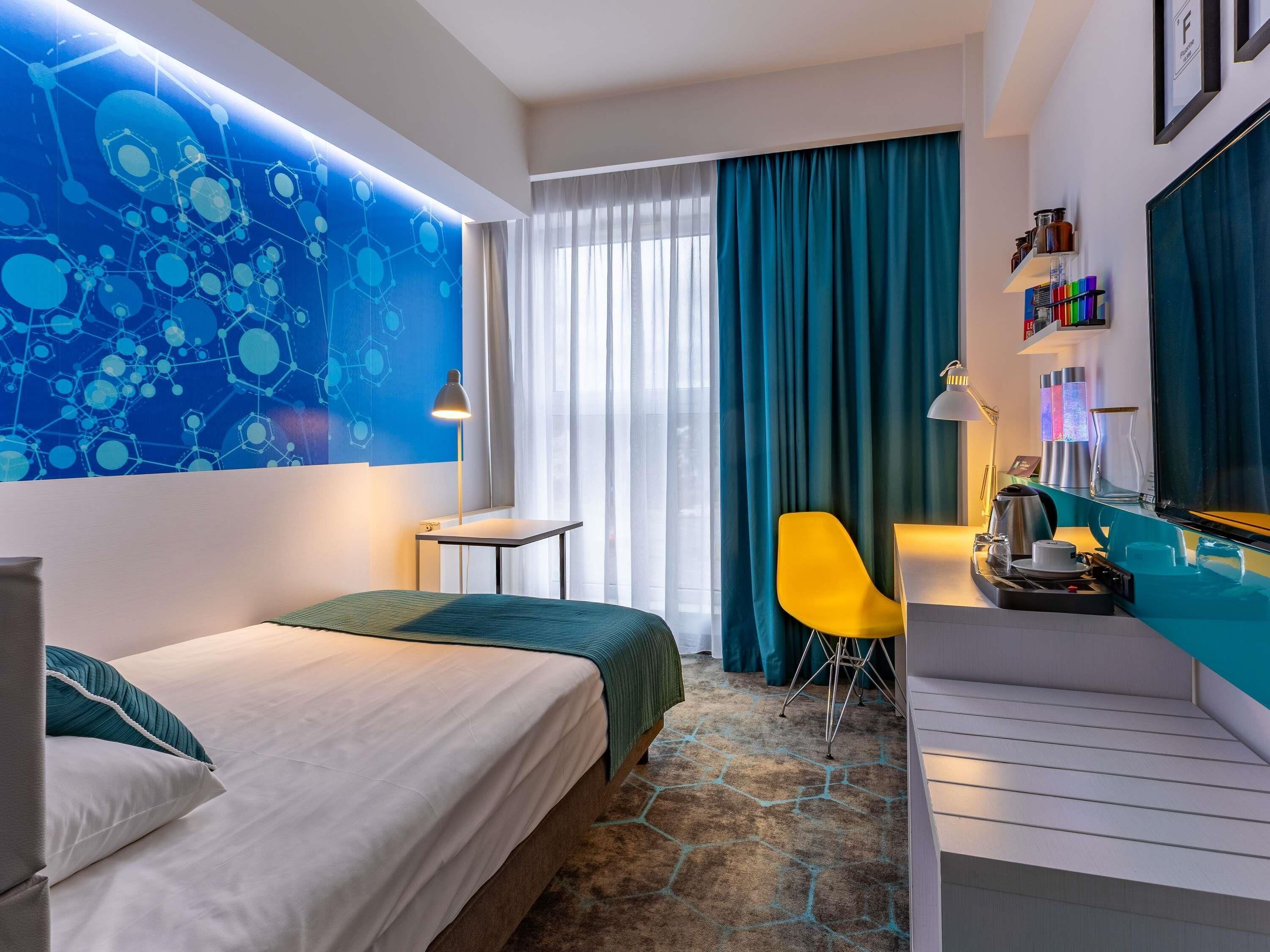 A hotel room with a bed, a desk, a chair, a large window with curtains, and a wall with a blue abstract design.