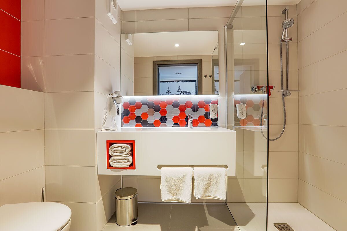 A modern bathroom with a glass shower enclosure, a white sink with a colorful hexagonal tile backsplash, and a small trash can.