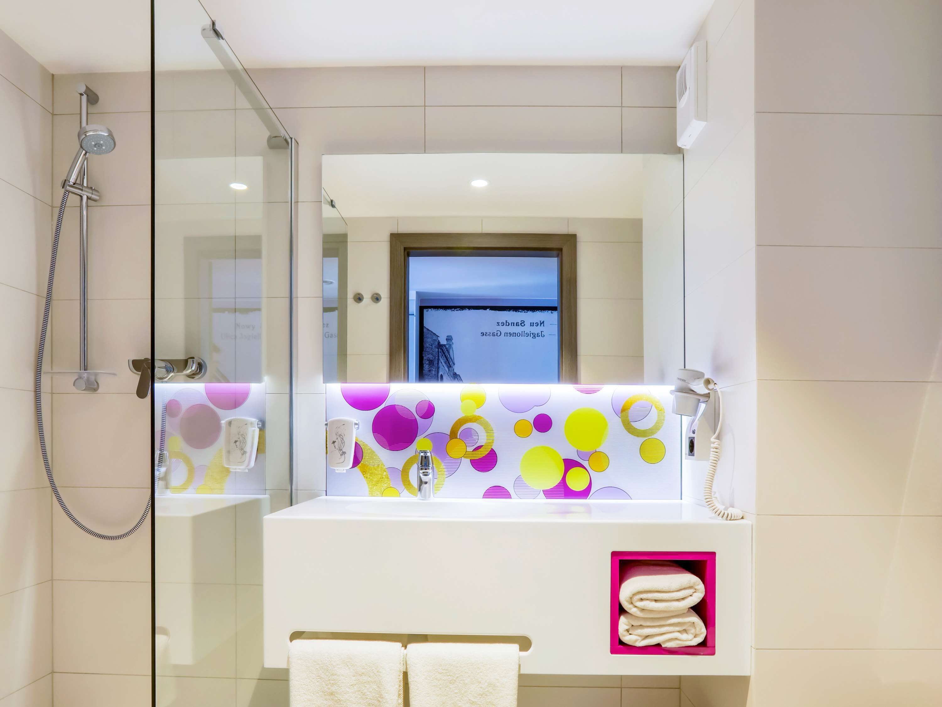 Casa de banho moderna com cabine de duche em vidro, lavatório branco com padrões circulares coloridos e espaço para toalhas em rosa.
