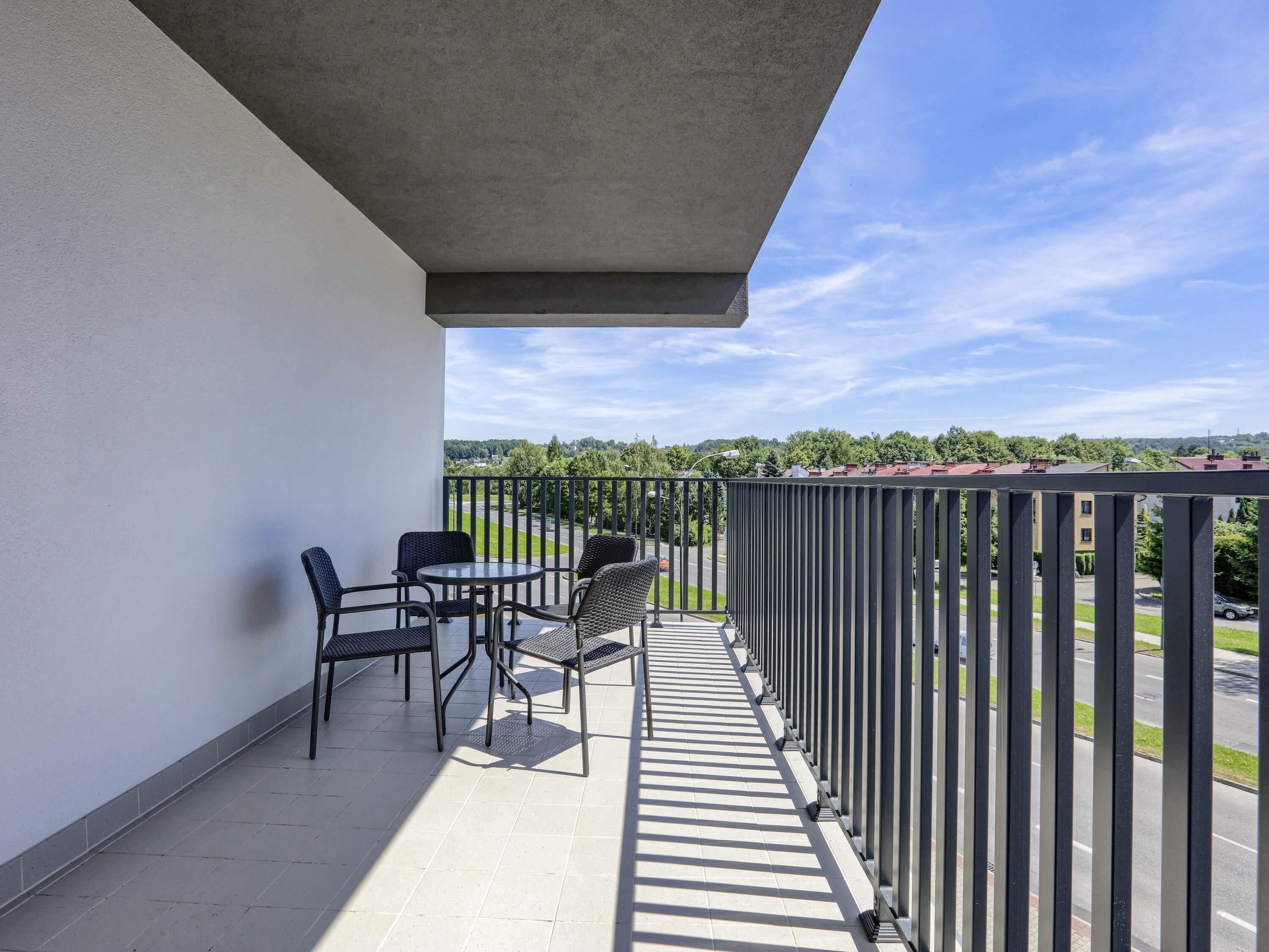 A balcony with a table and chairs, a view of a road and greenery, and a clear sky.