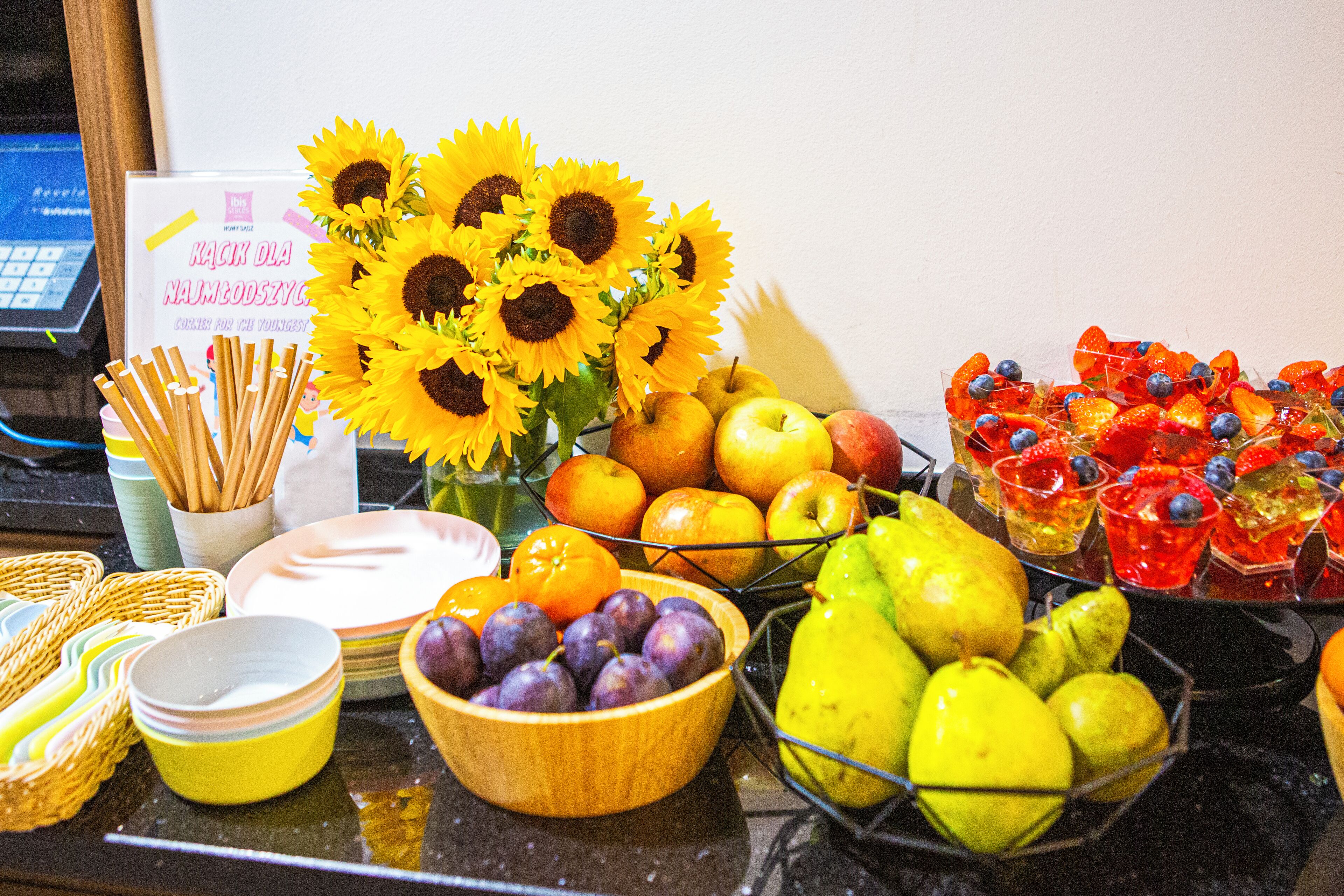 Una mesa con varias frutas, un cuenco de uvas, un jarrón de girasoles y un letrero en idioma extranjero.