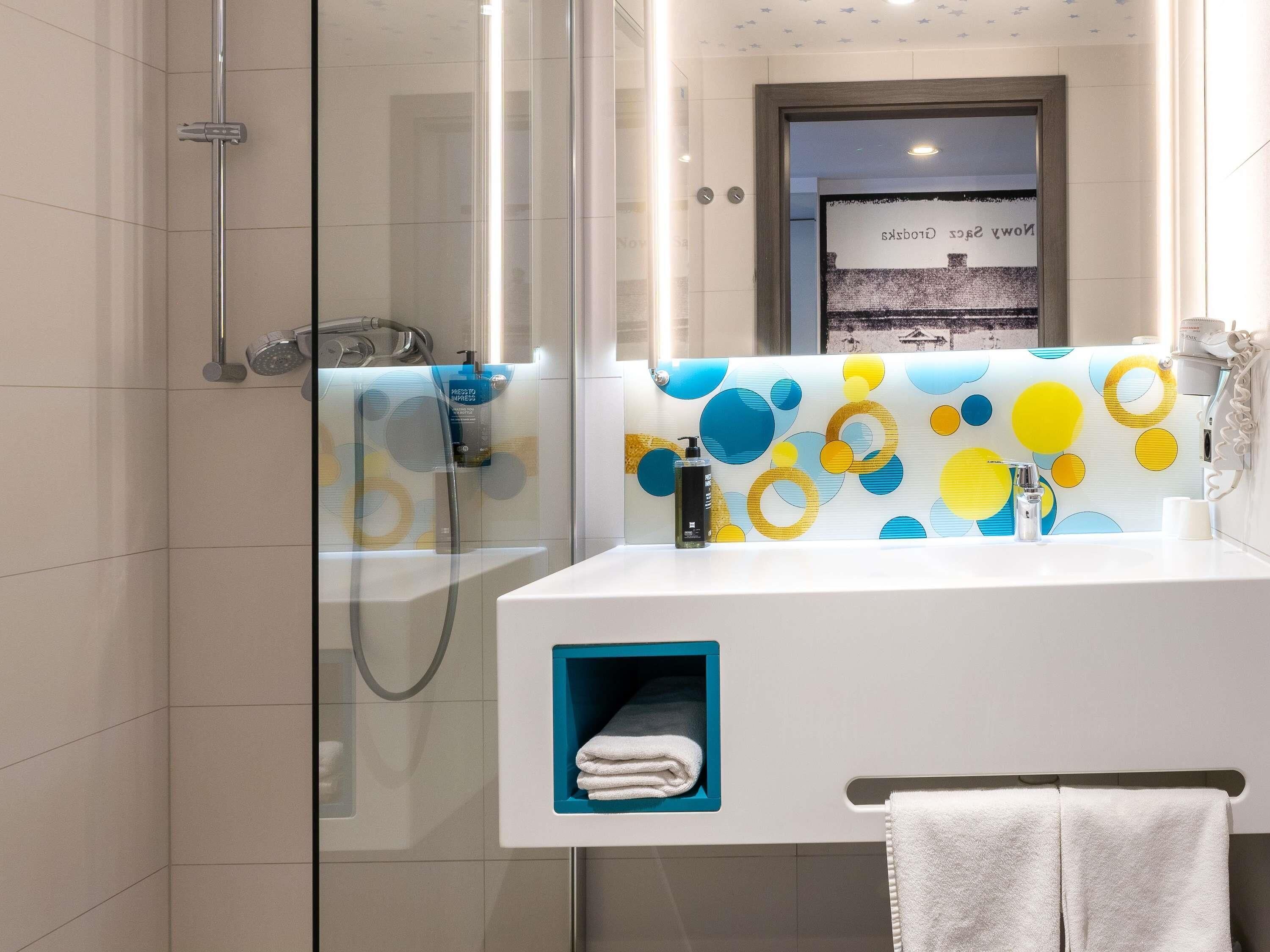 Une salle de bain moderne avec une douche à parois de verre, un lavabo blanc doté d’un distributeur de savon bleu et un porte-serviettes.