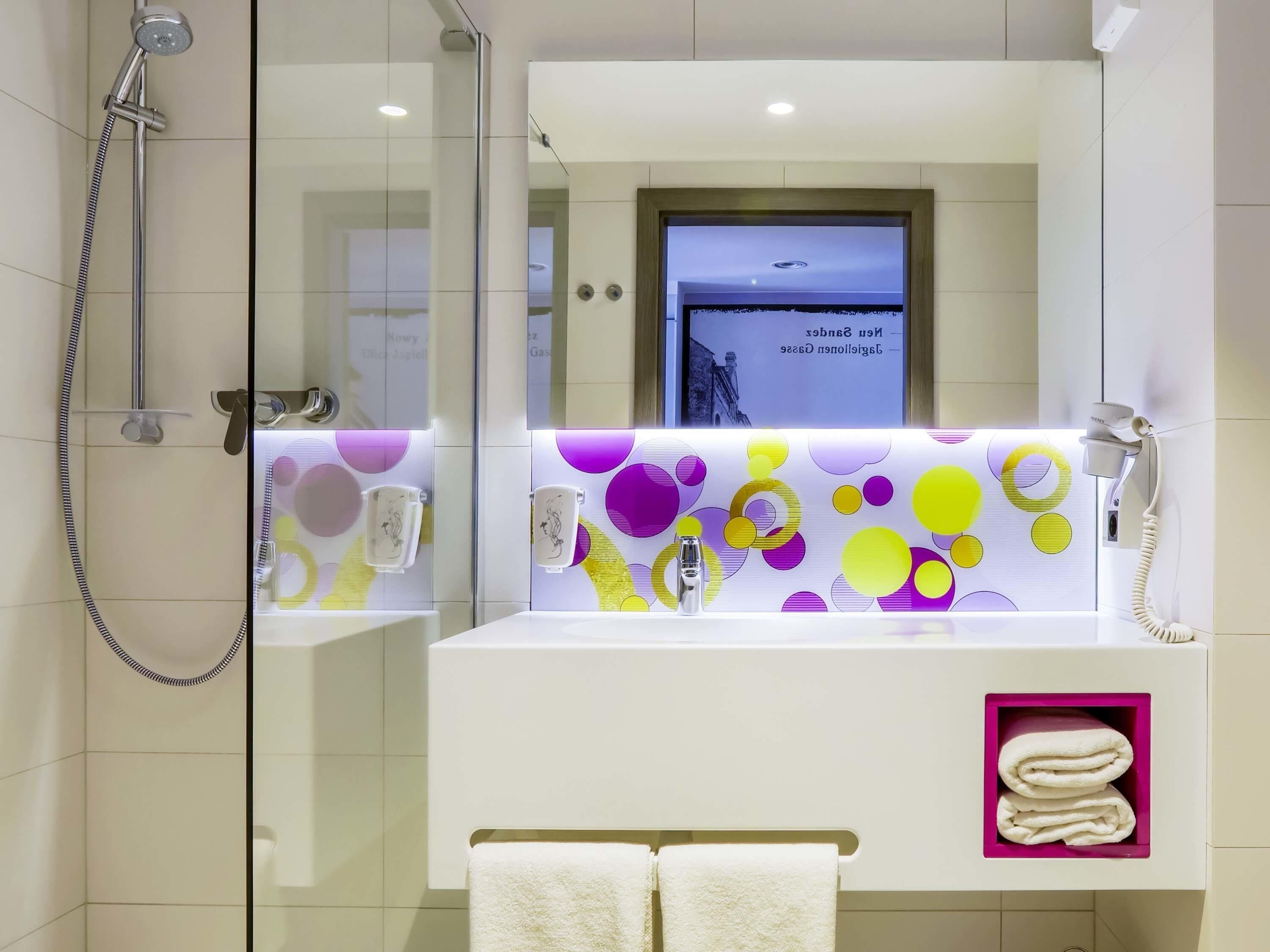 Un baño moderno con ducha de vidrio, lavamanos blanco y un salpicadero abstracto y colorido, además de un tocador con espejo.