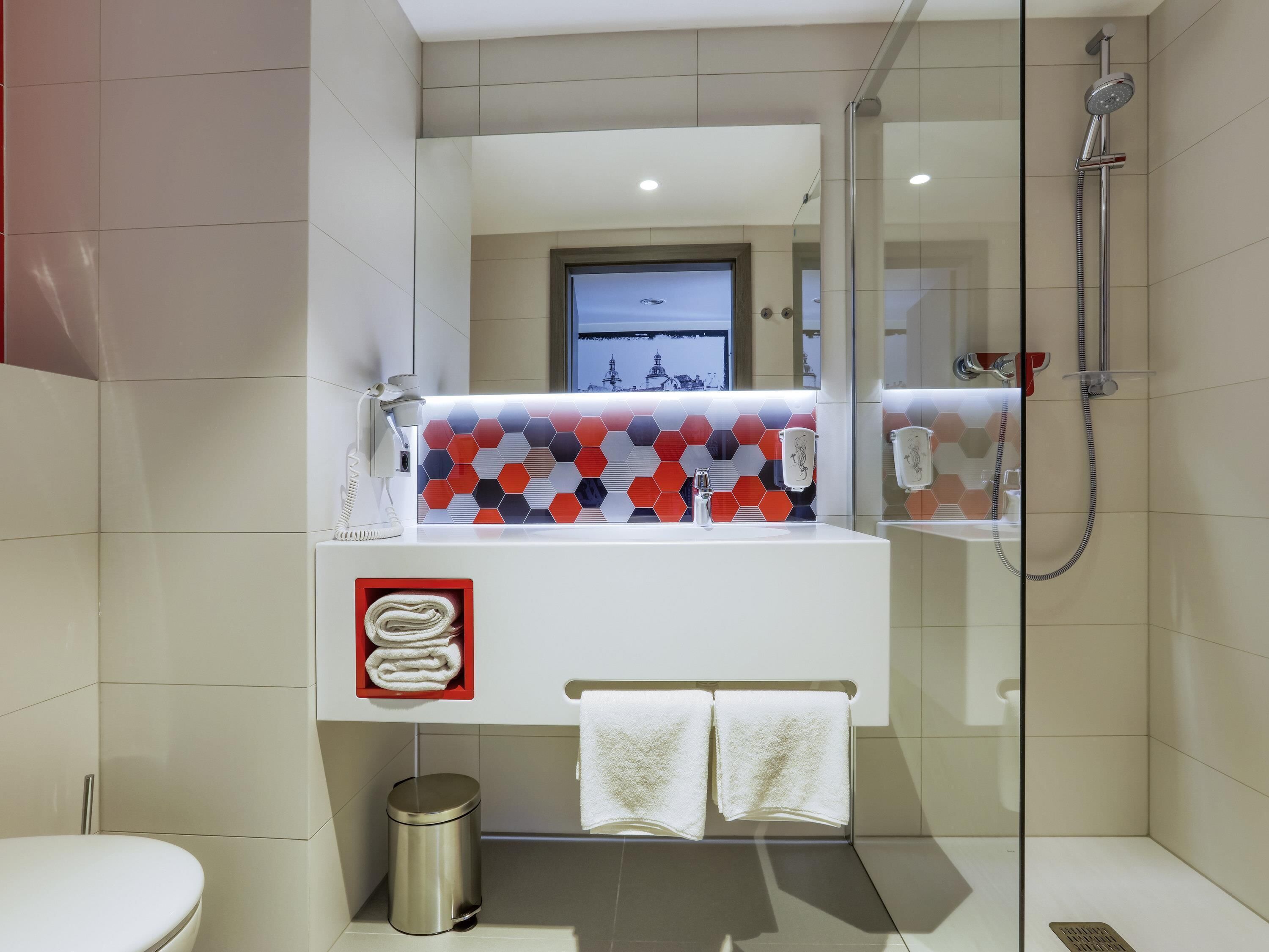 A modern bathroom with a glass-enclosed shower, a white sink with a colorful hexagonal backsplash, and a small trash bin.