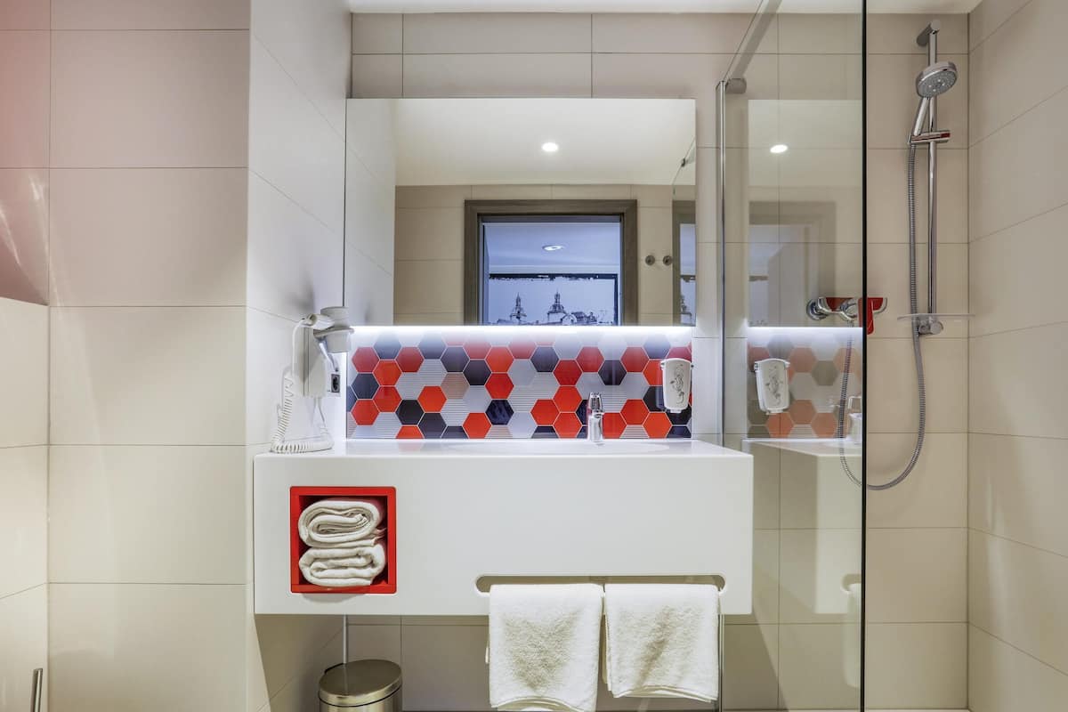 Un baño moderno con cabina de ducha de vidrio, un lavamanos blanco y un salpicadero de azulejos hexagonales rojo y negro, y una ventana visible.