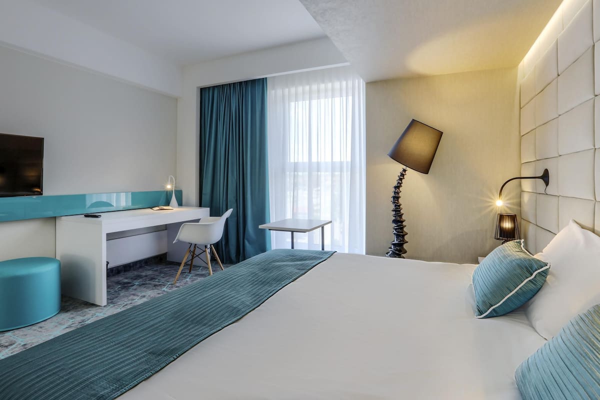 Una habitación de hotel moderna con una cama, un escritorio, una silla, un televisor, una lámpara y una ventana con cortinas.