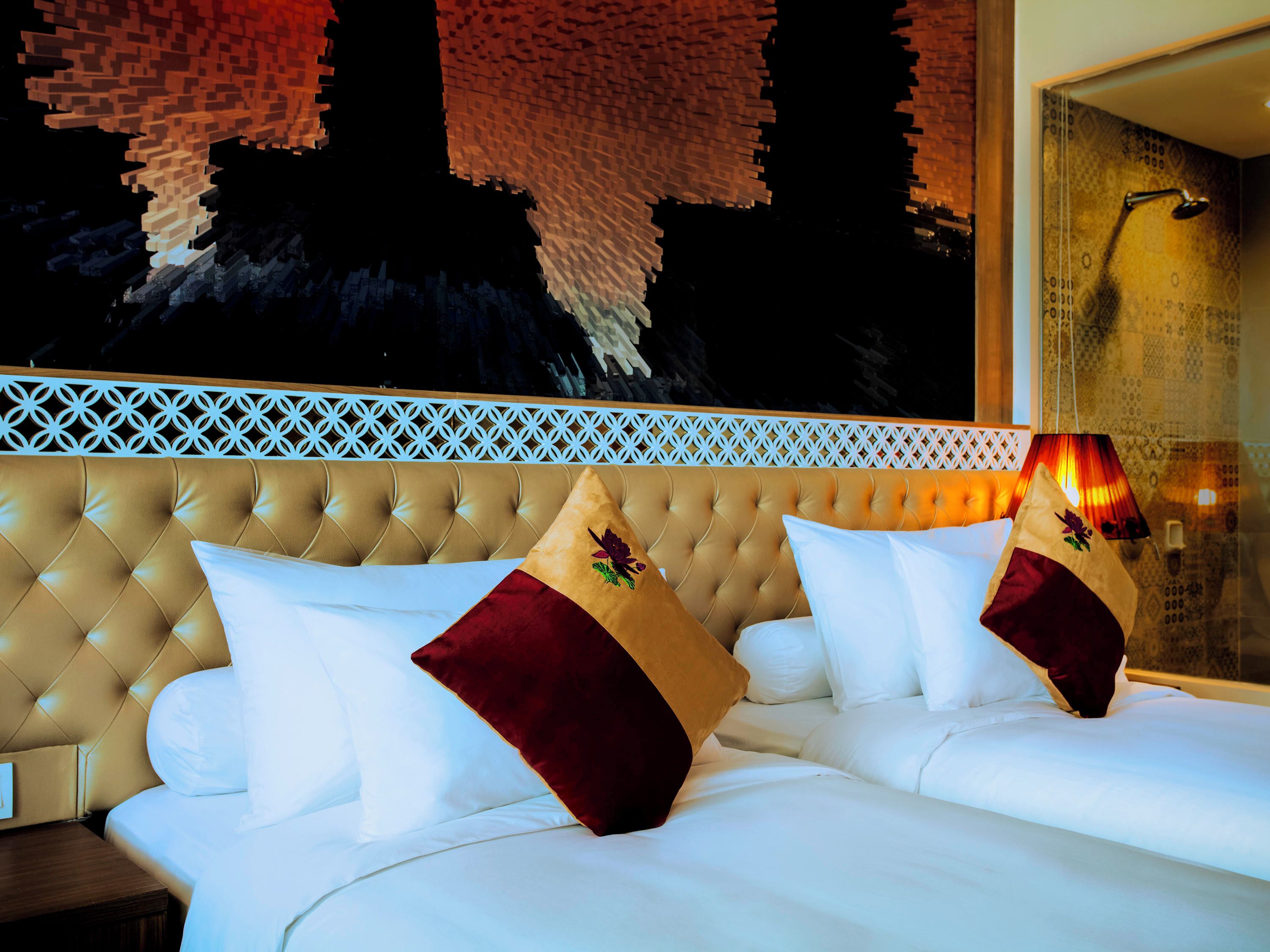 A hotel room with a tufted headboard, a bed with white linens and red and gold pillows, and a patterned wall.