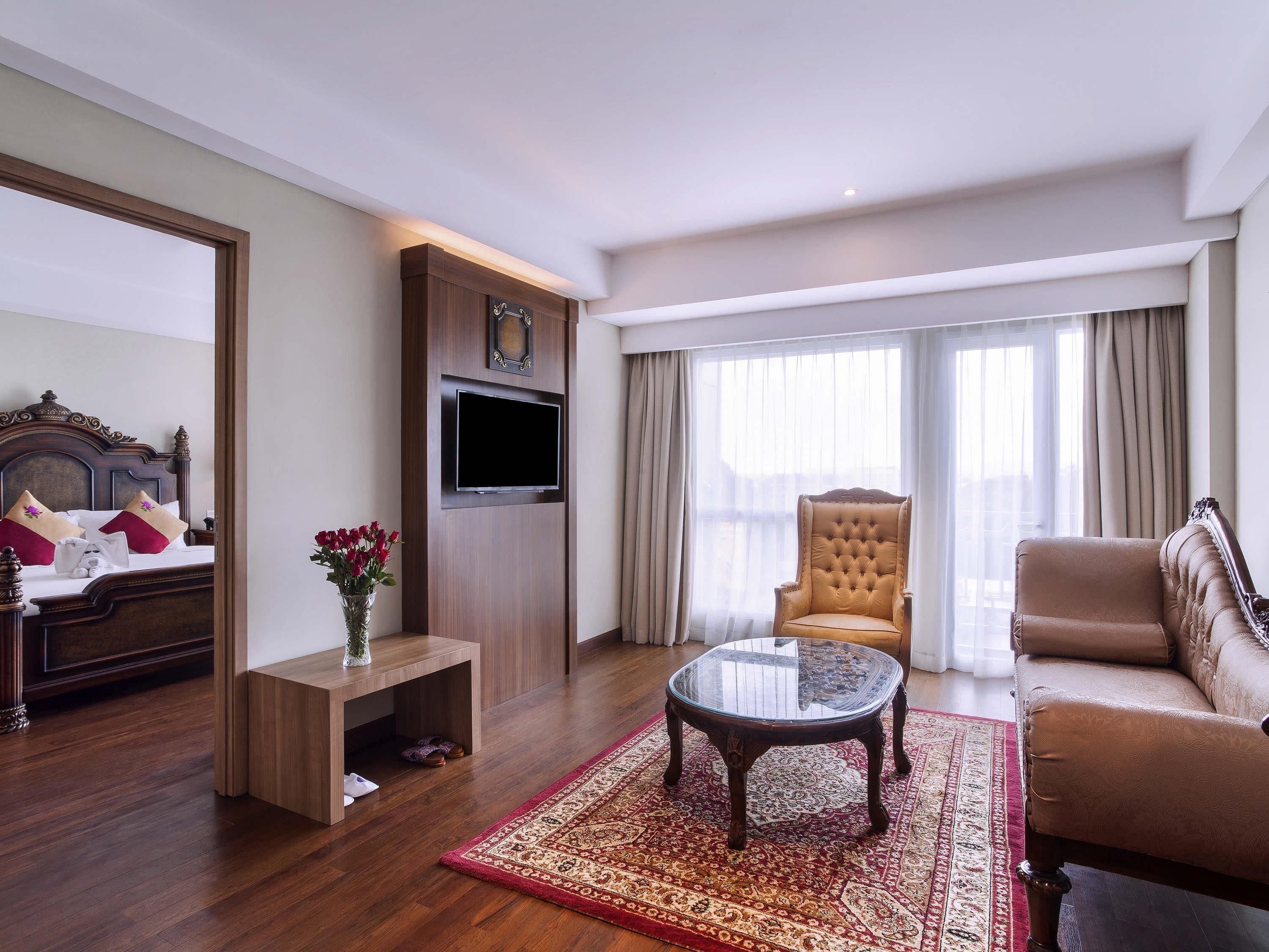 A hotel room with a bed, a TV mounted on a wooden panel, a leather armchair, a round coffee table, and a large window with curtains.