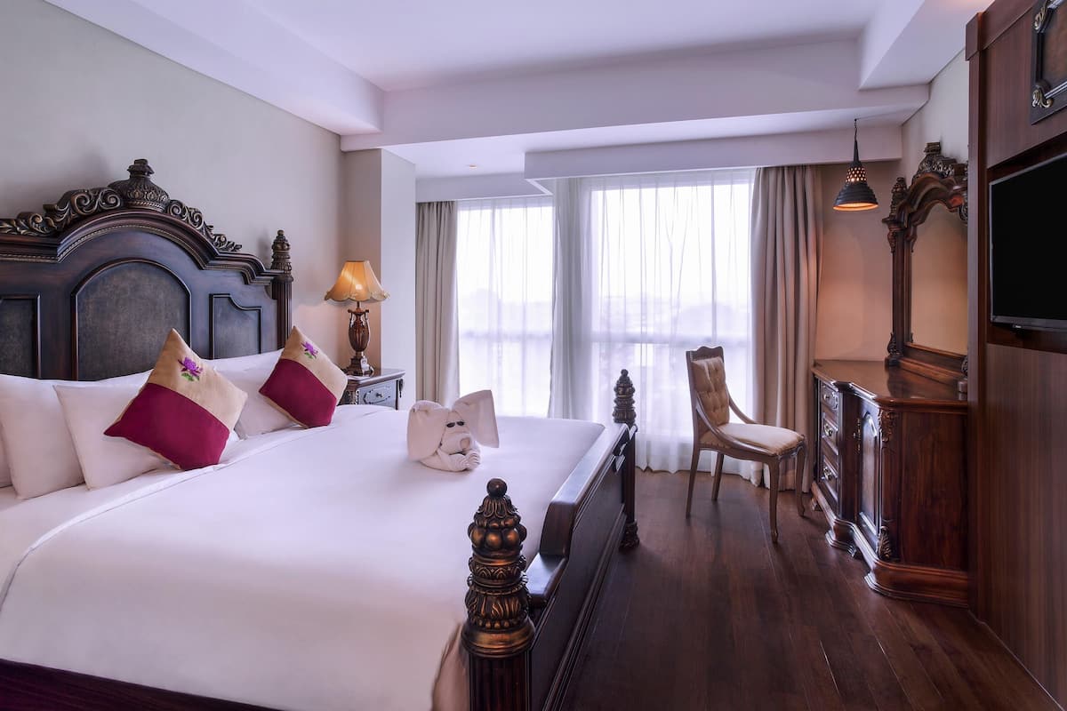 A hotel room with a large bed, a wooden headboard, a desk with a chair, a television, and a window with curtains.