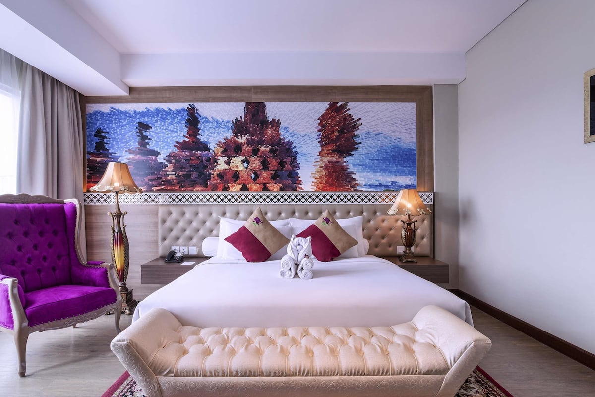 A hotel room with a large bed, a purple armchair, a decorative lamp, and a wall-mounted artwork.