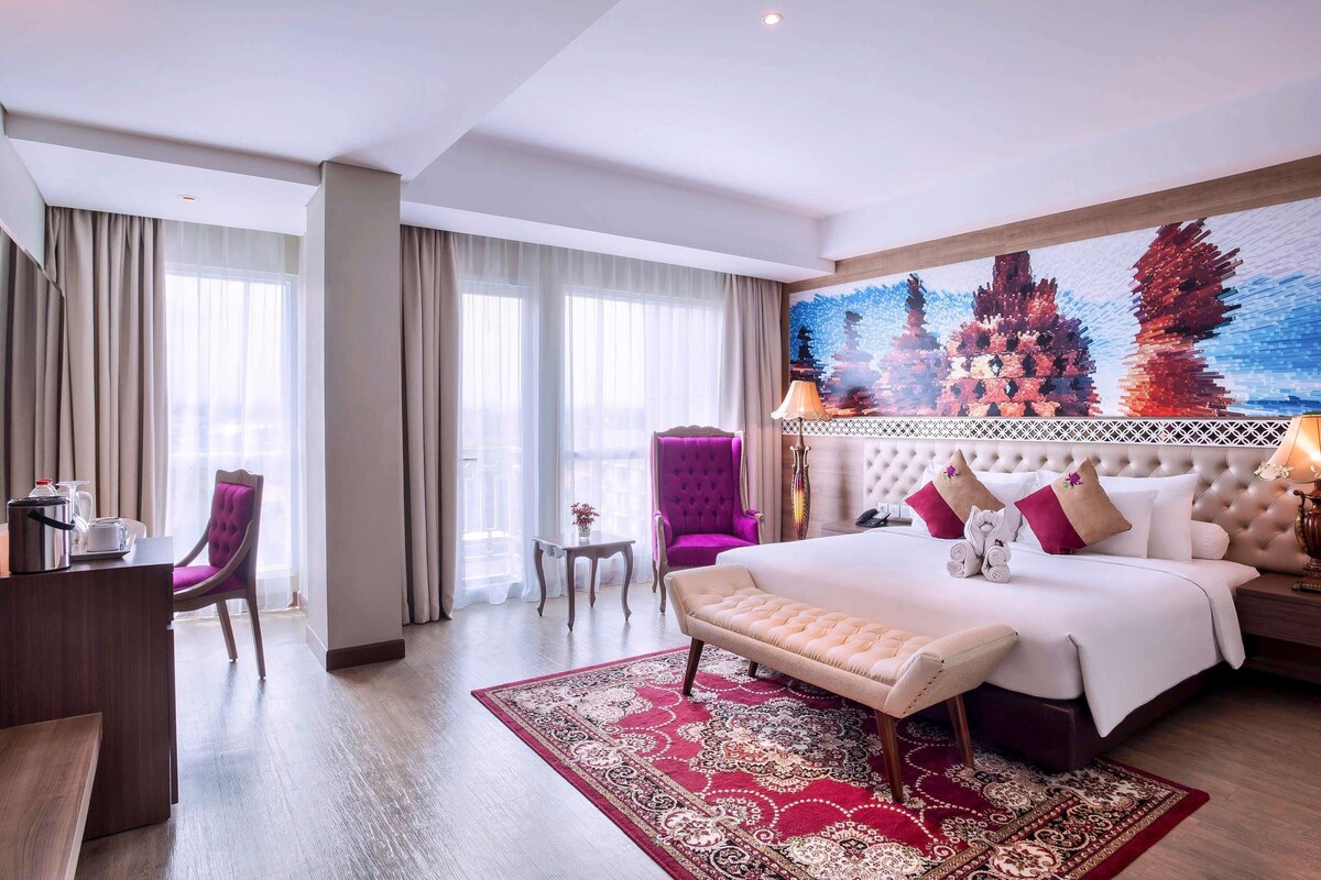 Ein Hotelzimmer mit Bett, Schreibtisch, Stuhl, kleinem Tisch und einem großen Fenster.
