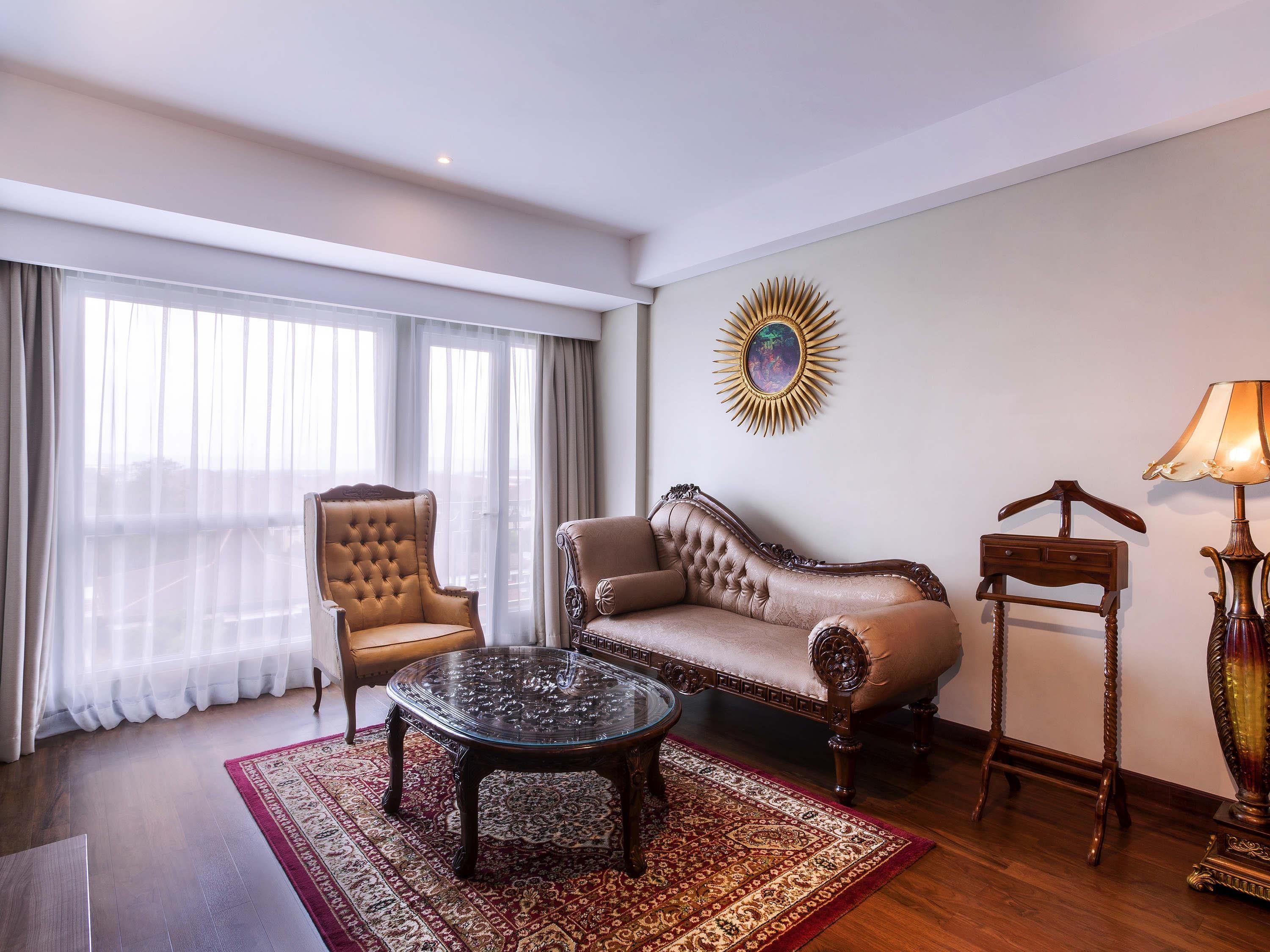 A hotel room with a large window, a sofa, a chair, a glass coffee table, a patterned rug, and a decorative wall piece.