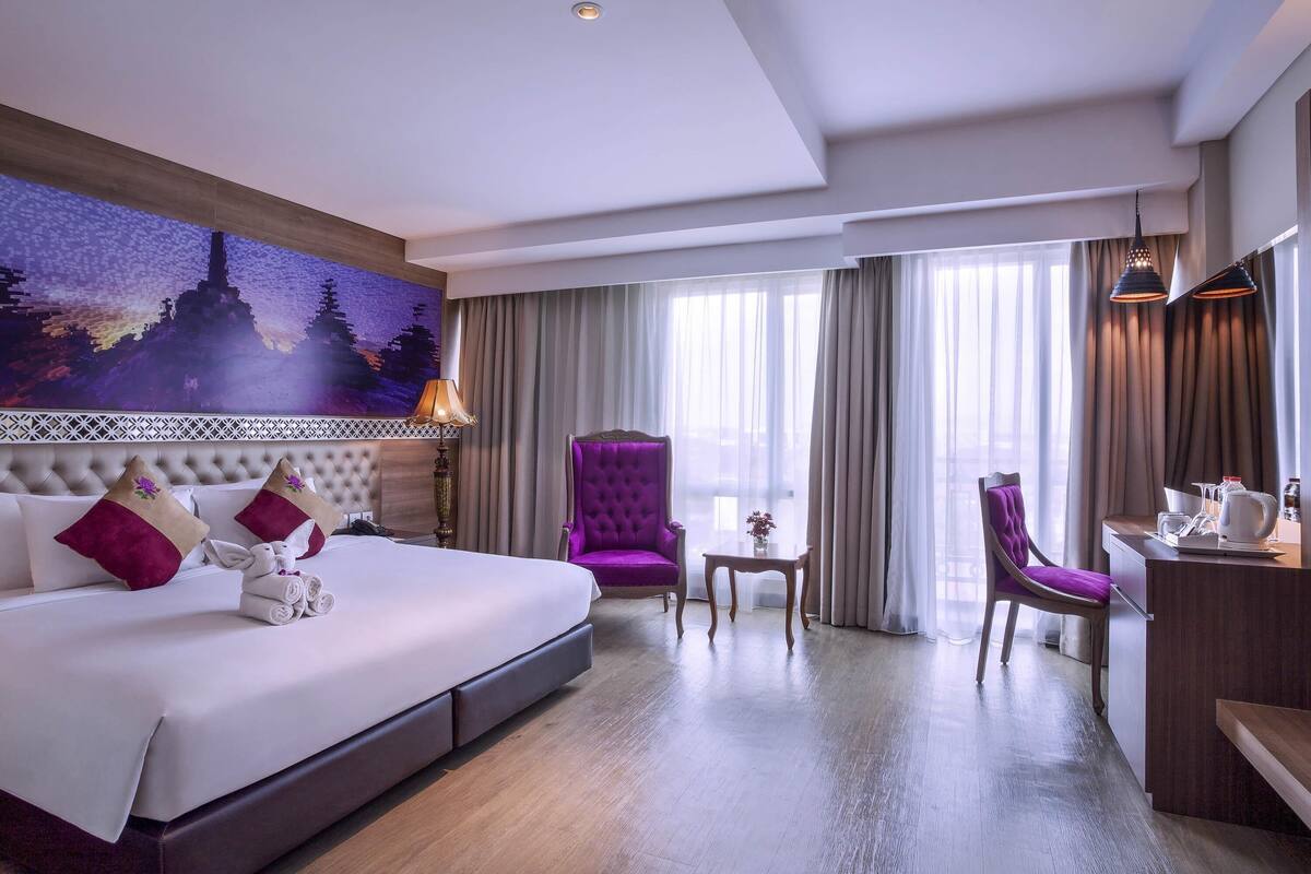 A hotel room with a large bed, a chair, a small table, and a desk with a coffee pot.