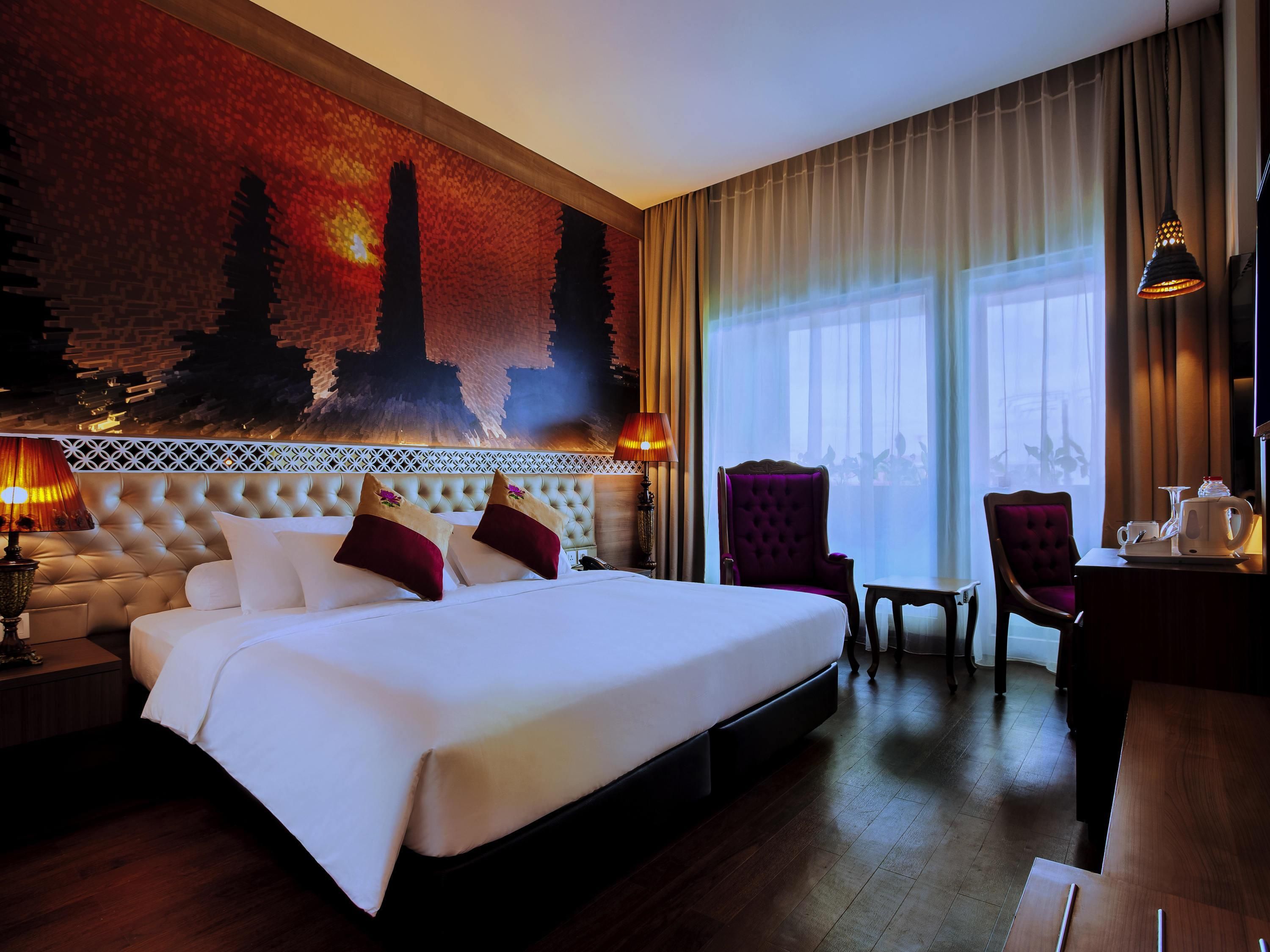 A hotel room with a large bed, two chairs, a small table with a kettle, and a view of a cityscape through the window.