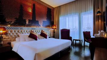 A hotel room with a large bed, two chairs, a small table with a kettle, and a view of a cityscape through the window.