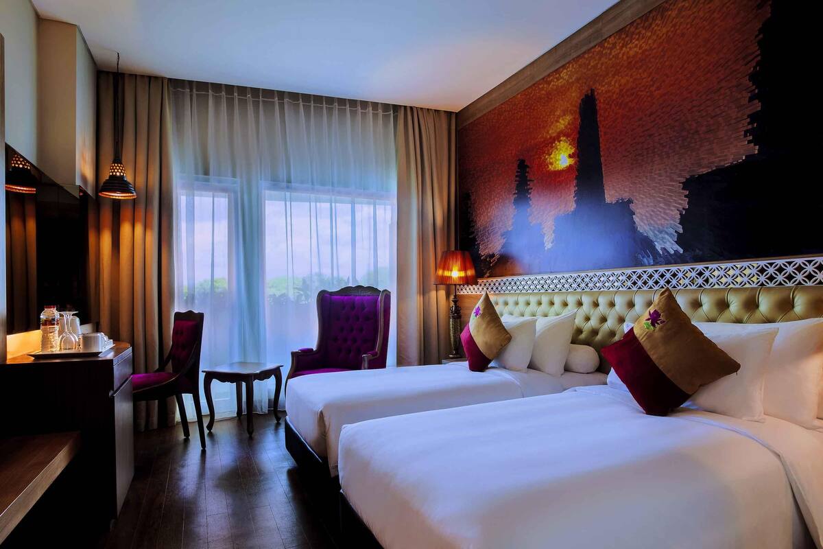 A hotel room with two beds, a desk with a chair, a table with a lamp, and a large window with curtains.
