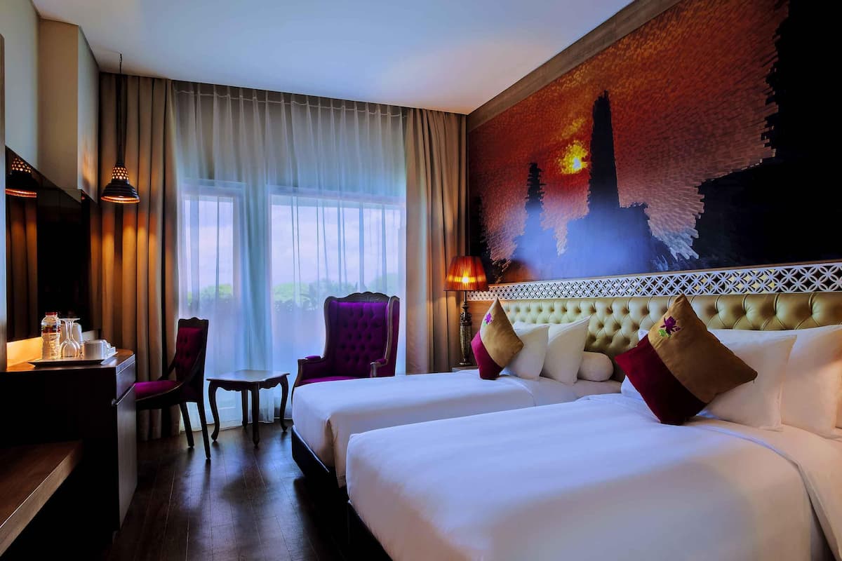 A hotel room with two beds, a desk with a chair, a table with a lamp, and a large window with curtains.