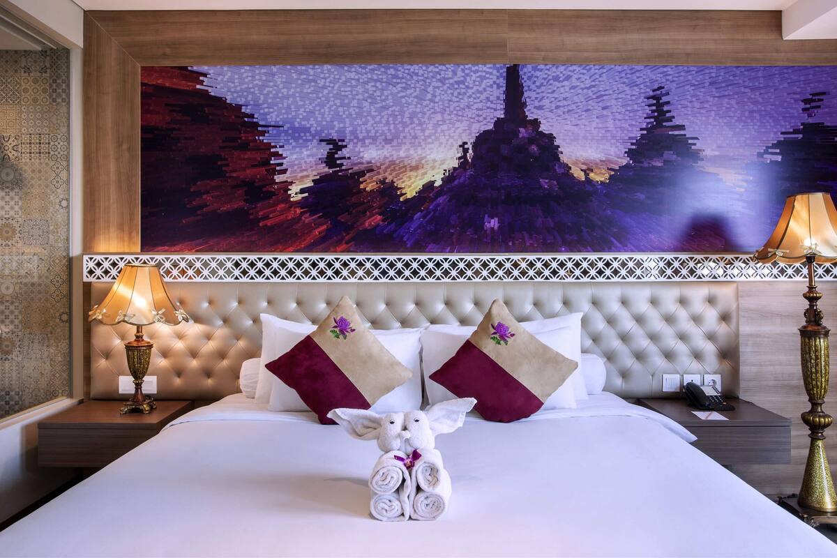 A hotel room with a large bed, a decorative headboard, a wall-mounted artwork, a nightstand with a lamp, and a telephone.