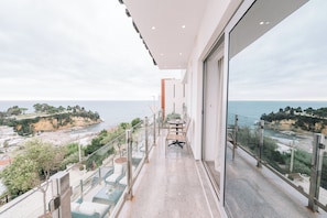 Un balcón con barandilla de vidrio que ofrece vistas al mar, una mesa con sillas y un cielo despejado.