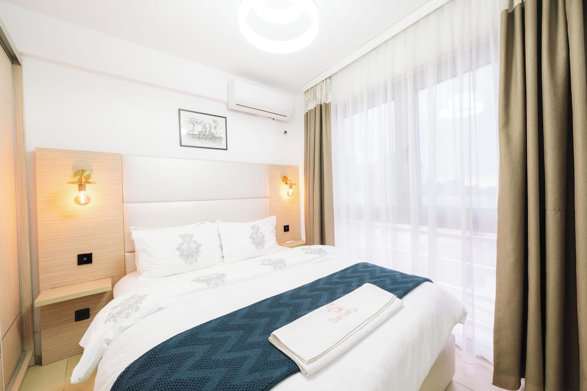 Una habitación de hotel con una cama grande, dos mesitas de noche, un aire acondicionado de pared y una ventana con cortinas traslúcidas.