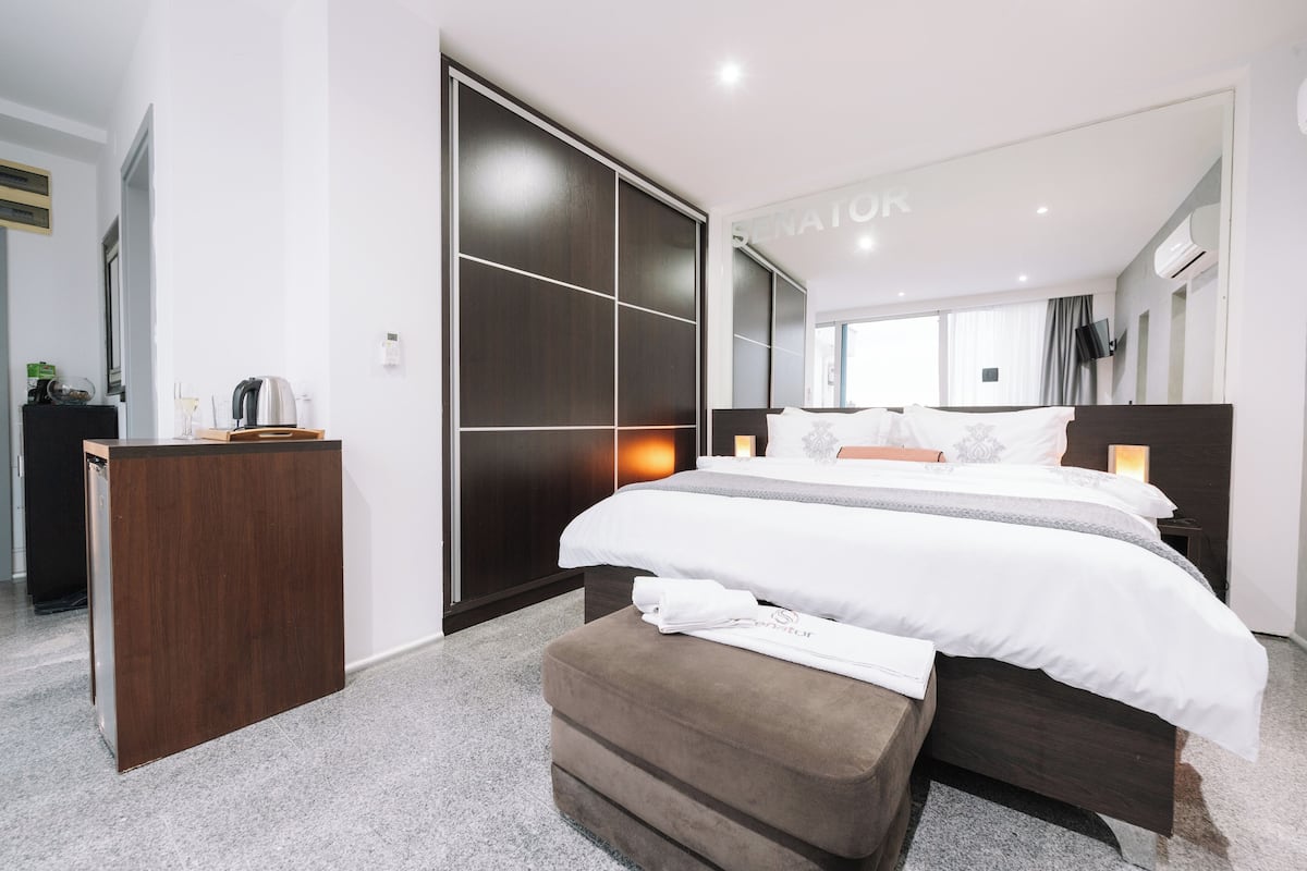 Una habitación de hotel moderna con una cama grande, un armario de madera, un escritorio pequeño y un espejo en la pared.