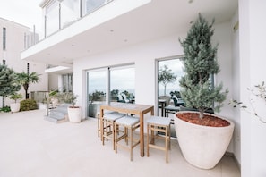 Un patio moderno al aire libre con una mesa y sillas de madera, plantas grandes en macetas y vistas de la ciudad a través de amplias ventanas.