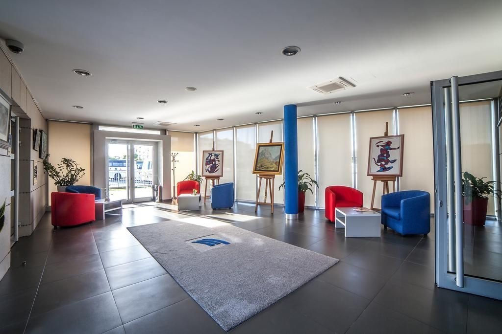 A modern lobby with a grey carpet, red and blue chairs, and paintings on easels.